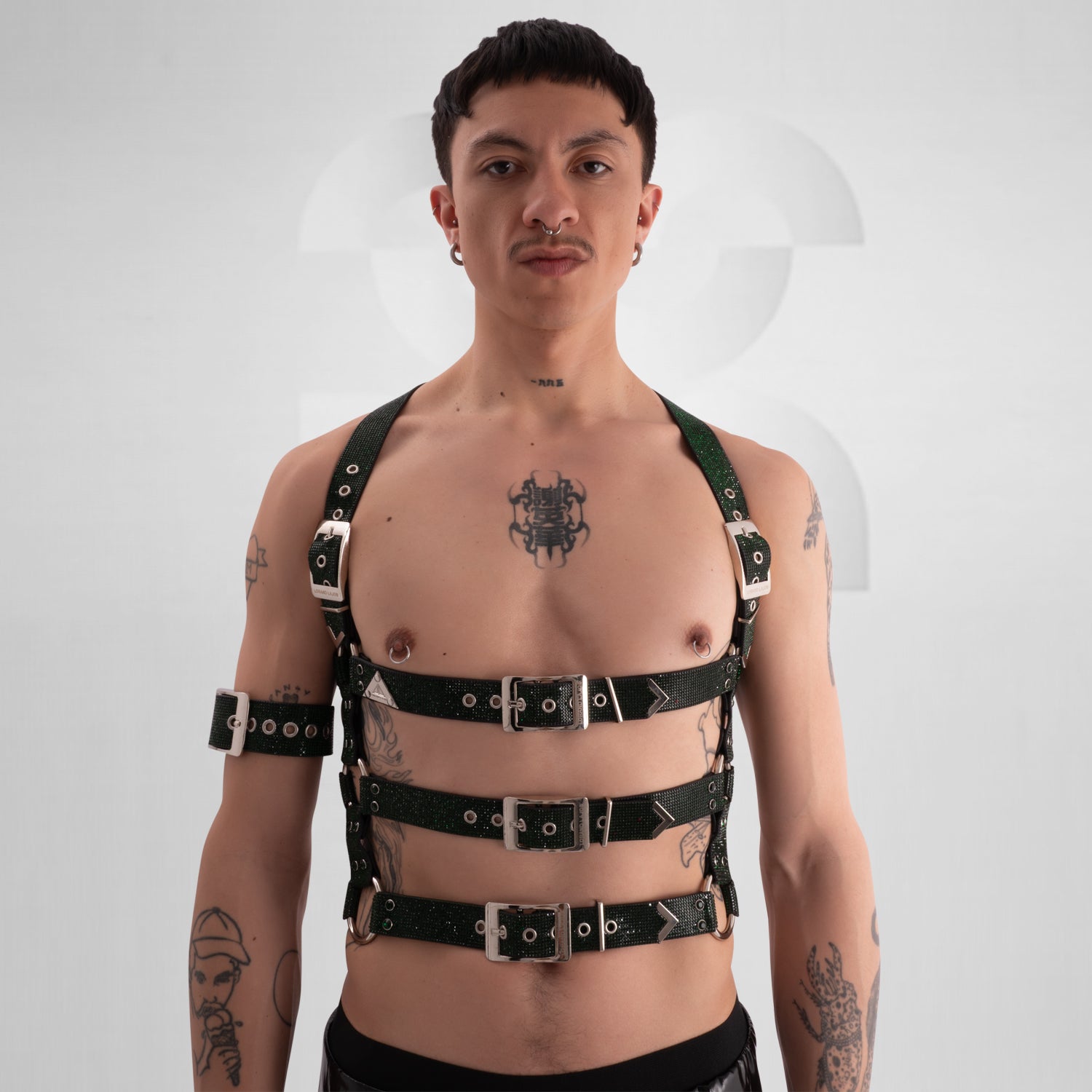 This image showcases a person with a confidently rebellious style, featuring multiple tattoos on their chest, arms, and neck. They have short, dark hair and a septum piercing. The individual is wearing a bold, black harness made of sturdy, glittering material with multiple adjustable straps and silver hardware, which encases the chest and shoulders. Their makeup is minimal, emphasizing their natural features. The background is plain with subtle geometric shapes, putting full focus on their striking and unco
