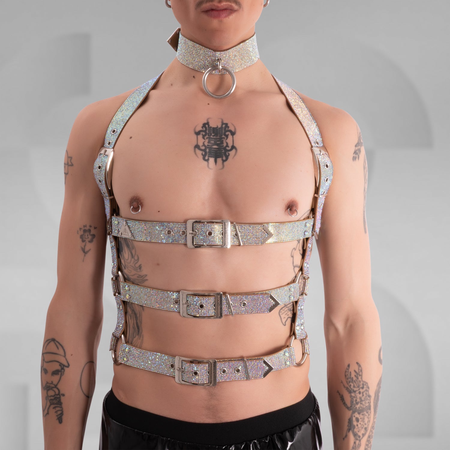 This image showcases a person with a confidently rebellious style, featuring multiple tattoos on their chest, arms, and neck. They have short, dark hair and a septum piercing. The individual is wearing a bold, black harness made of sturdy, glittering material with multiple adjustable straps and silver hardware, which encases the chest and shoulders. Their makeup is minimal, emphasizing their natural features. 