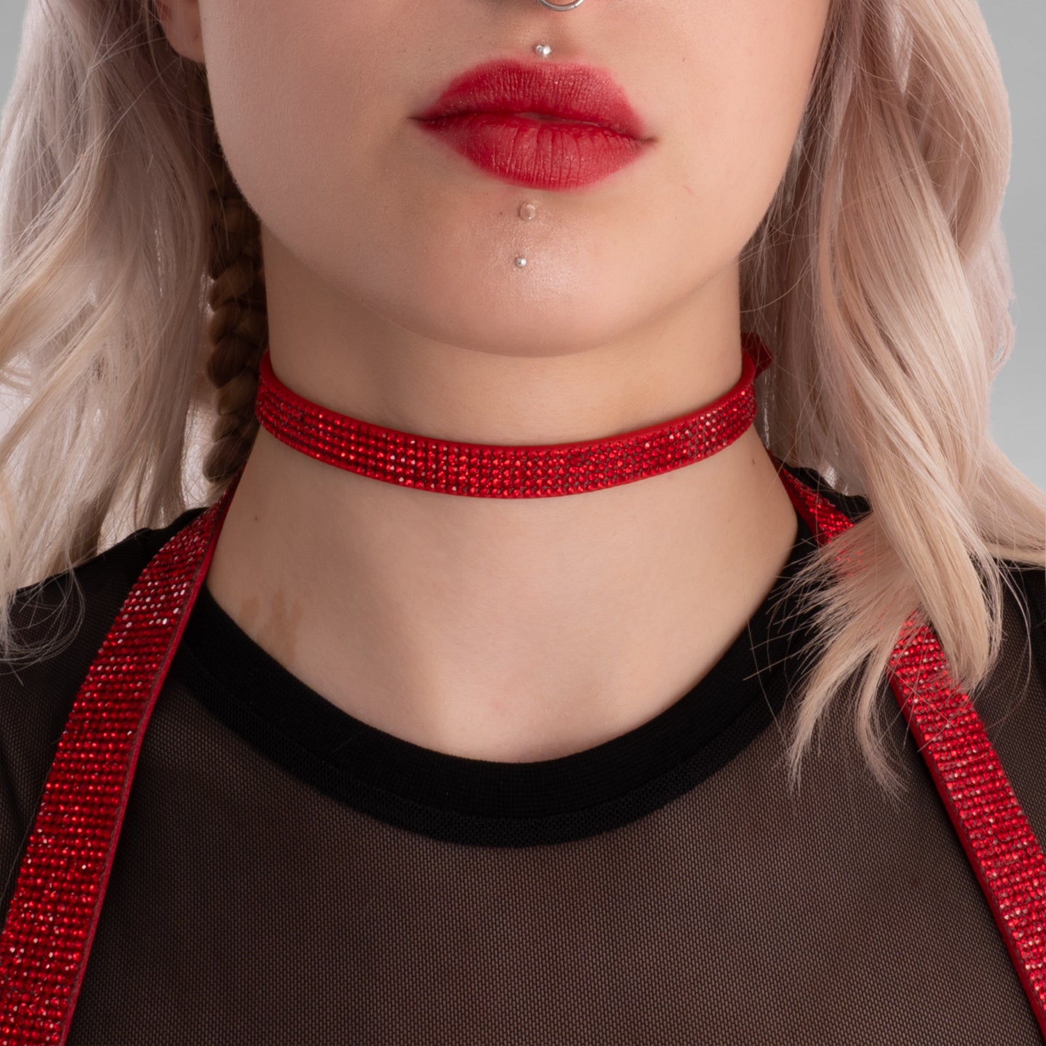 In this picture, you can see a close-up of a woman wearing a striking red rhinestone choker around her neck, which adds a glamorous and edgy vibe. She has platinum blonde hair styled with soft waves, and a small braid on one side. Her makeup features bold red lipstick that matches her choker, and she has a septum piercing as well as two dermal piercings below her lips, adding to her alternative, fashionable style. She is dressed in a black, sheer top with a round neckline, which complements her edgy accesso