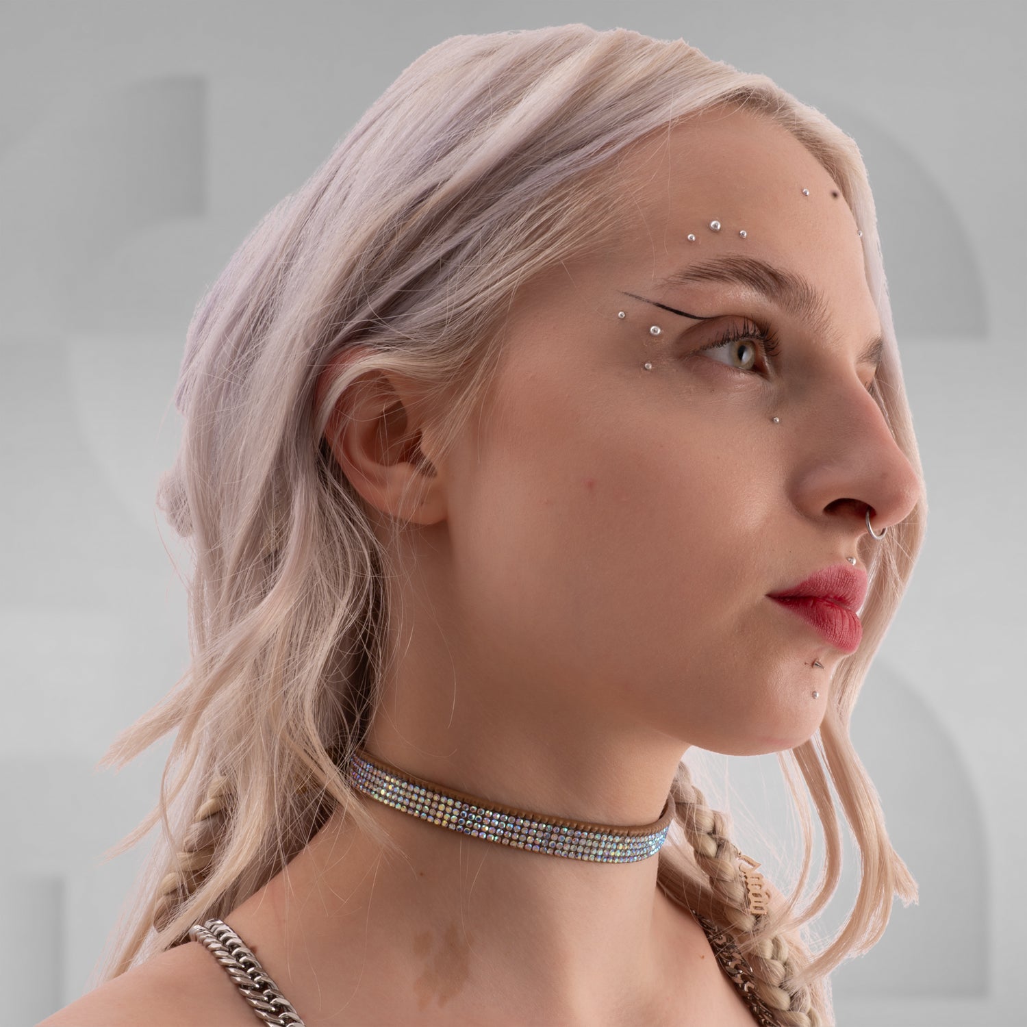 In this picture, you can see a close-up of a woman wearing a striking white rhinestone choker around her neck, which adds a glamorous and edgy vibe. She has platinum blonde hair styled with soft waves, and a small braid on one side. Her makeup features bold red lipstick that matches her choker, and she has a septum piercing as well as two dermal piercings below her lips, adding to her alternative, fashionable style. 
