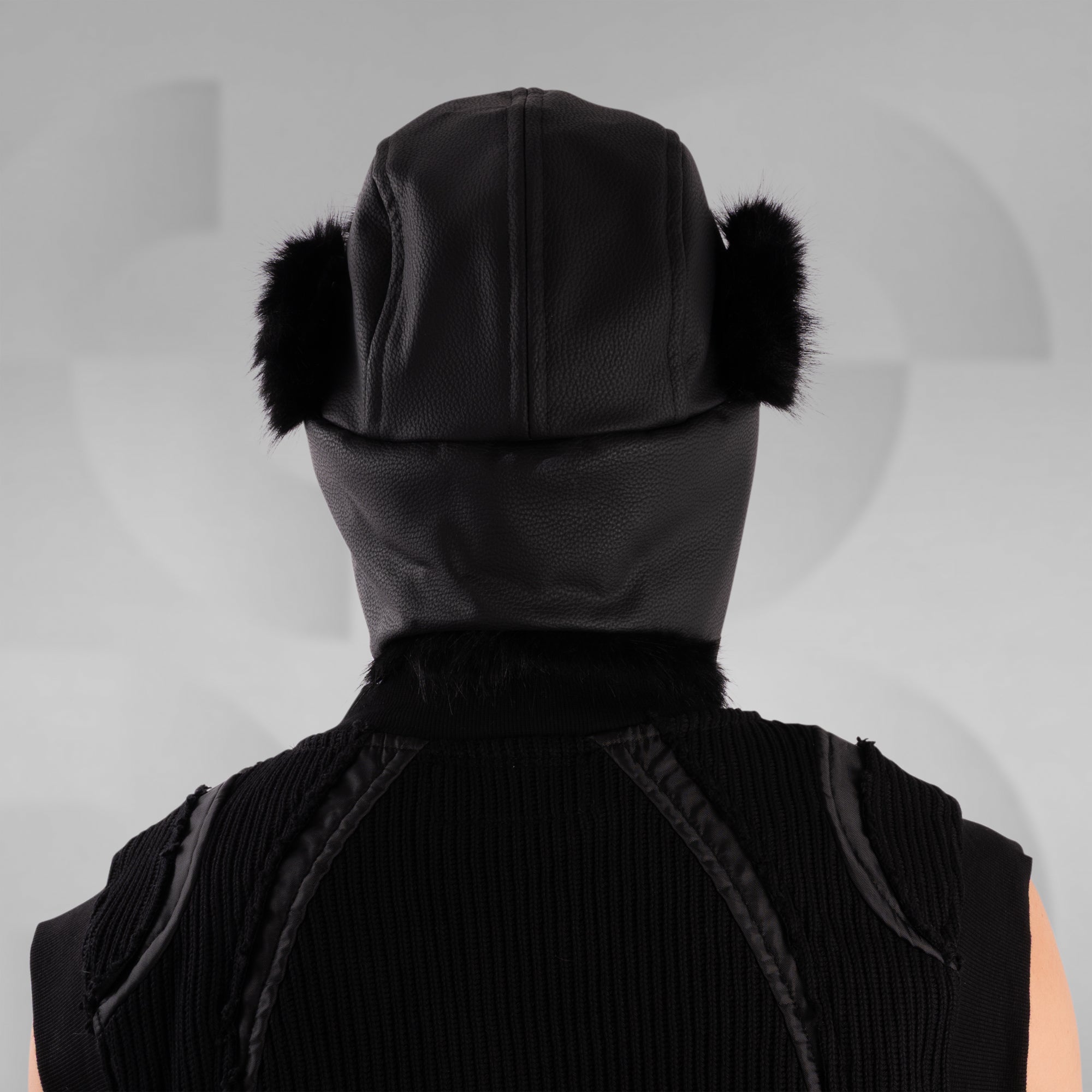 Person wearing a black hooded outfit with fur ear muffs on a gray background