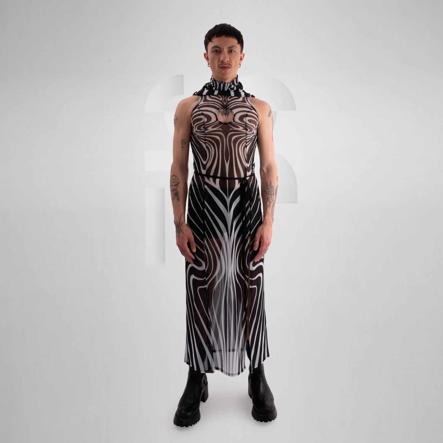 Model wearing a sleeveless, sheer black and white optical illusion print mesh dress with a high neck and cut-out chest detail, styled with black combat boots, standing confidently against a minimalist grey background everything looks very tribal festival like.