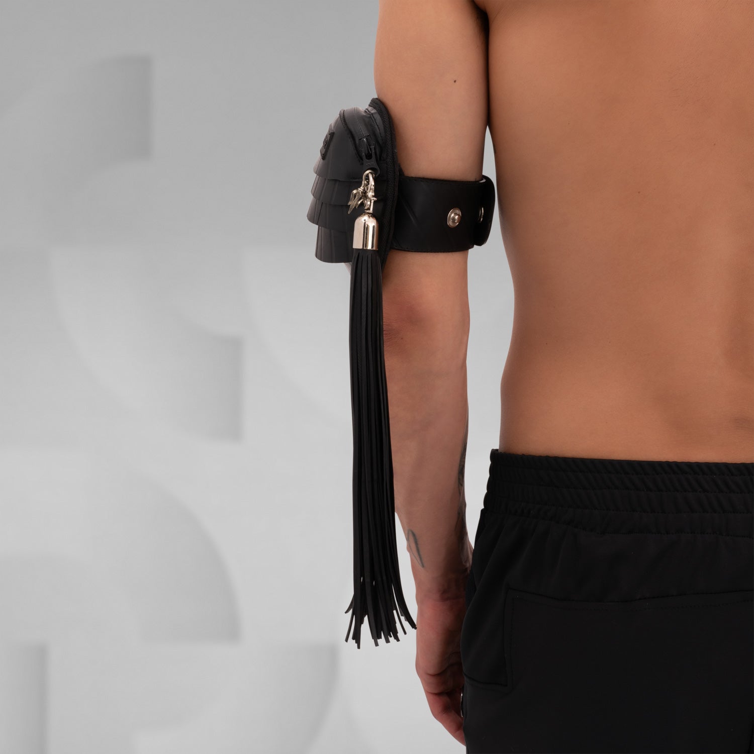 Side view of a black rubber whip accessory with metal clasp, attached to an arm pouch, designed to be clipped onto a fan or other accessories for added flair in clubwear.