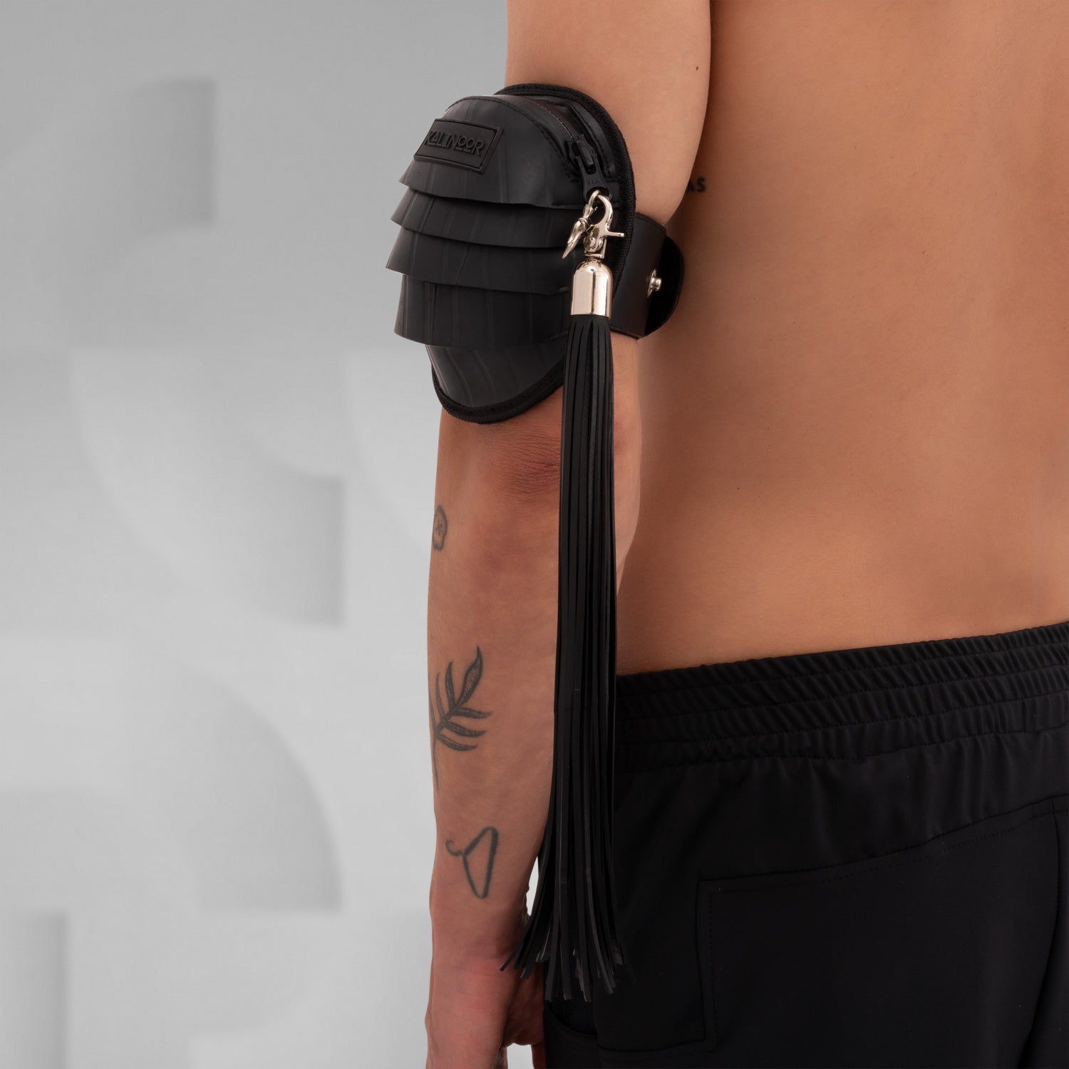 Side view of a black rubber whip accessory with metal clasp, attached to an arm pouch, designed to be clipped onto a fan or other accessories for added flair in clubwear.