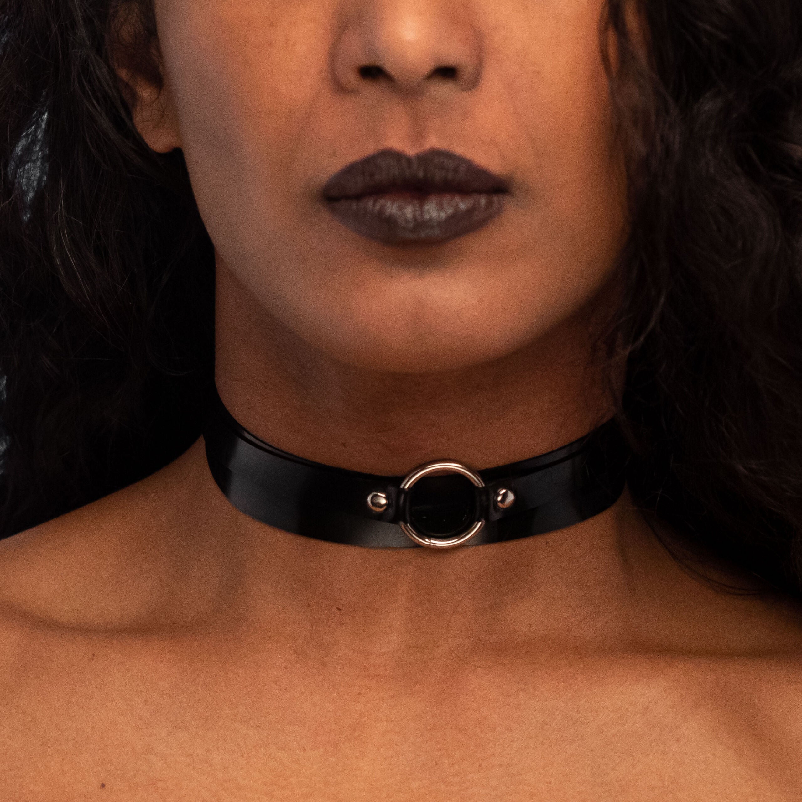 The image features a person showcasing a striking black latex choker, elegantly hugging the neck. The choker features a central metal ring that adds a bold detail to the overall design. The model's dramatic makeup, including vibrant eye shadow and dark lips, complements the edgy aesthetic of the accessory, making it a perfect statement piece for any outfit. This look is ideal for nightlife, enabling the wearer to express confidence and style.