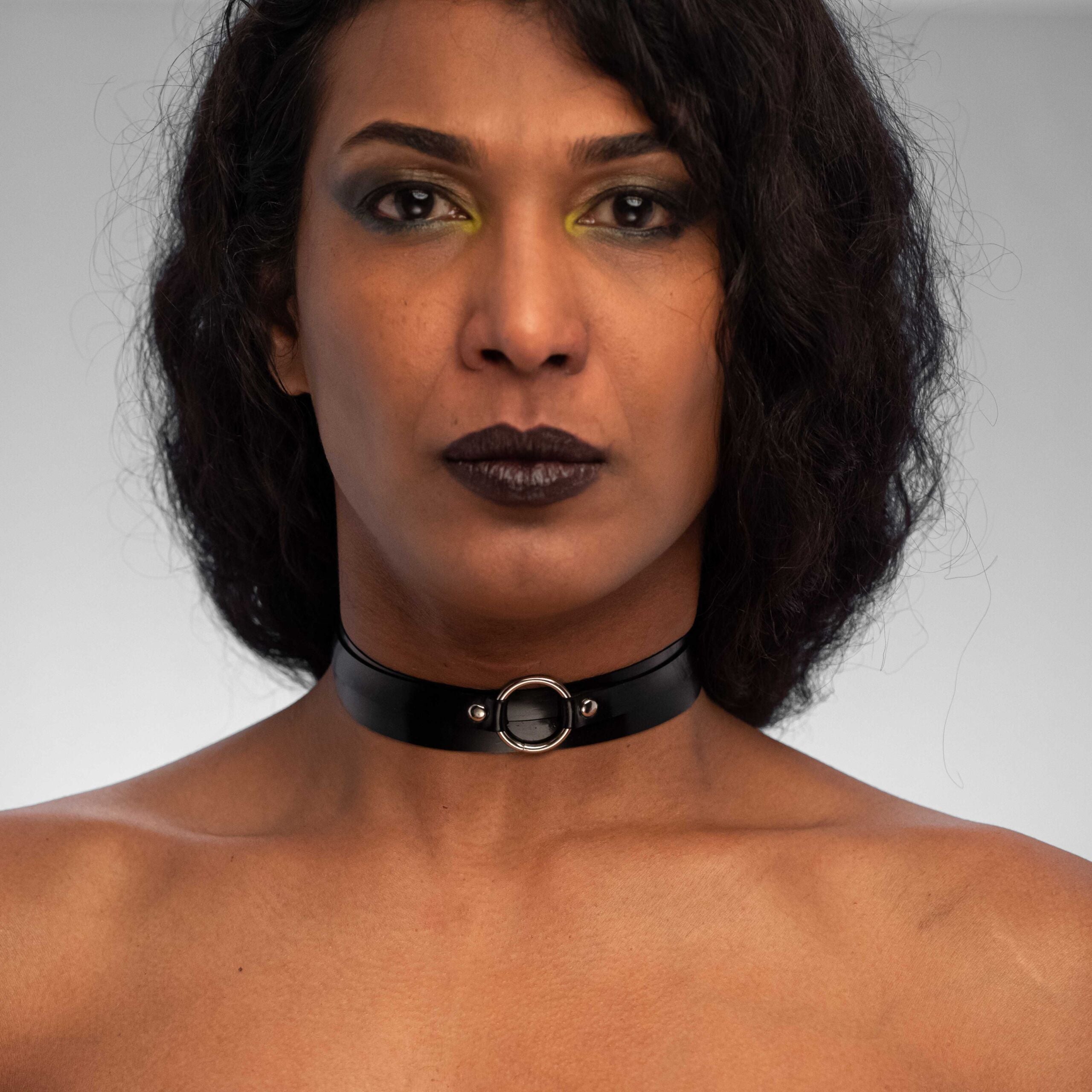 The image features a person showcasing a striking black latex choker, elegantly hugging the neck. The choker features a central metal ring that adds a bold detail to the overall design. The model's dramatic makeup, including vibrant eye shadow and dark lips, complements the edgy aesthetic of the accessory, making it a perfect statement piece for any outfit. This look is ideal for nightlife, enabling the wearer to express confidence and style.