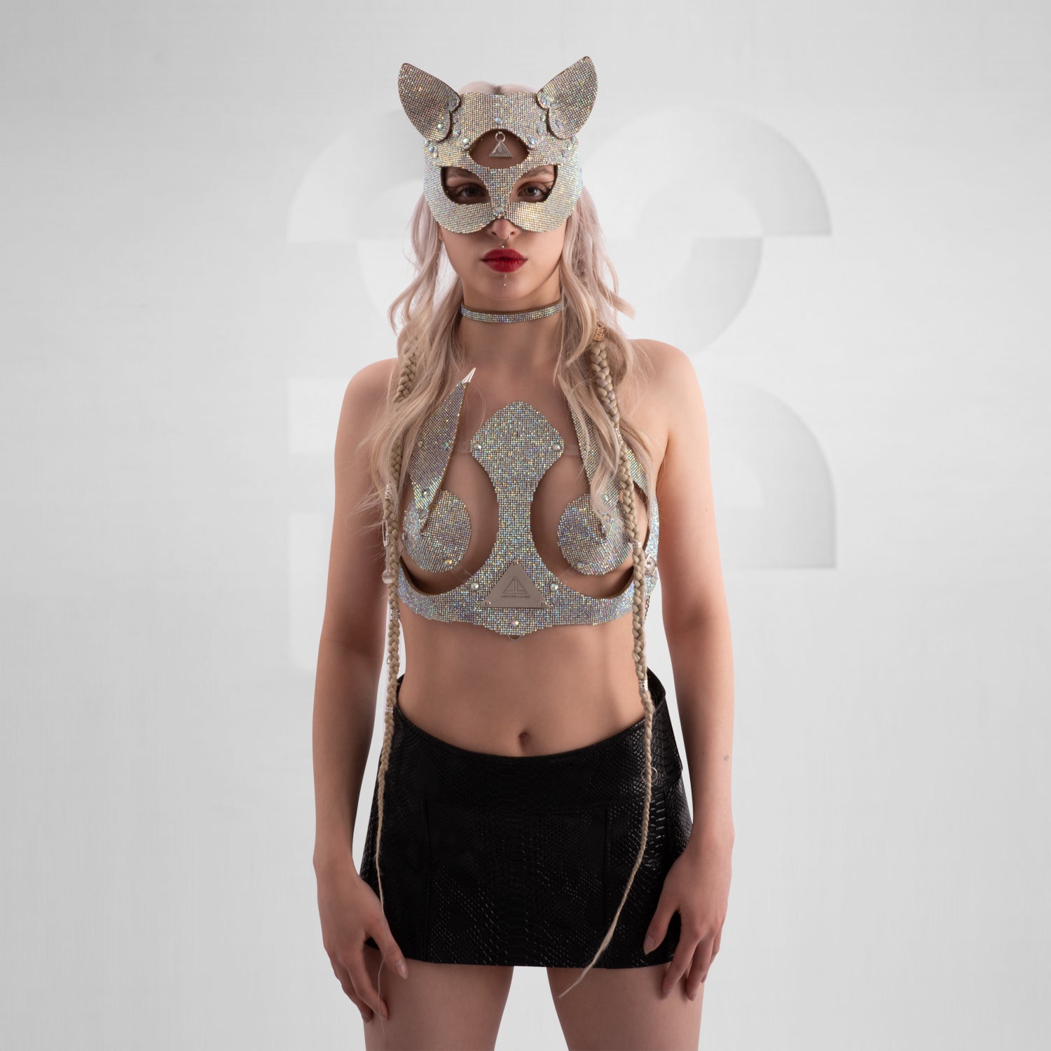 Woman wearing a sparkling rhinestone cat mask with pointed ears, paired with a matching rhinestone choker and shimmering outfit; styled with platinum blonde hair in braids and bold red lipstick, set against a minimalist grey background.