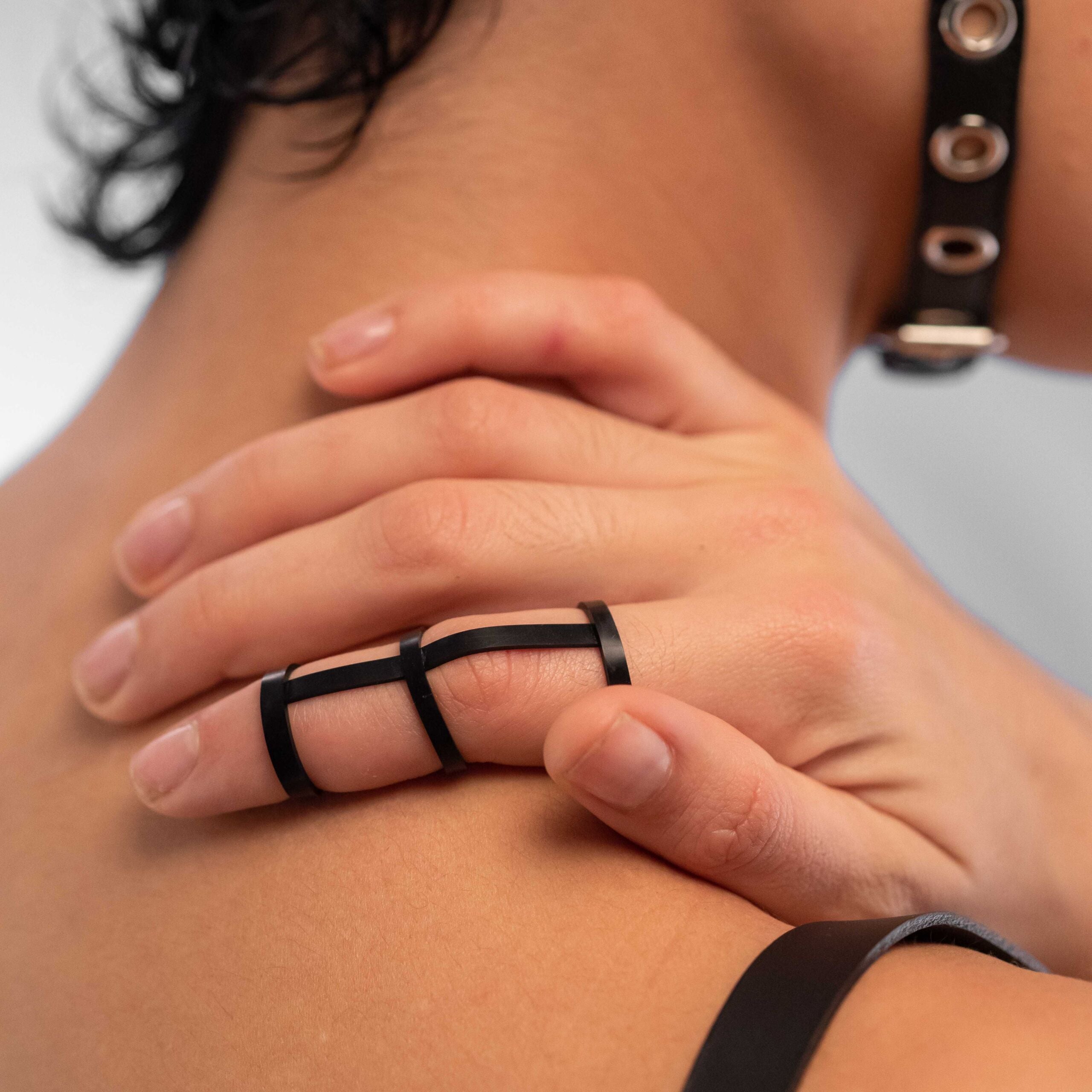 The image showcases a close-up of a hand wearing a sleek black latex finger harness, elegantly wrapping around the fingers and complementing the skin. This striking accessory highlights a modern, edgy aesthetic, seamlessly blending with the wearer’s style. The soft curves and minimalist design add a bold touch, making it perfect for both creative outfits and daring statements at parties.