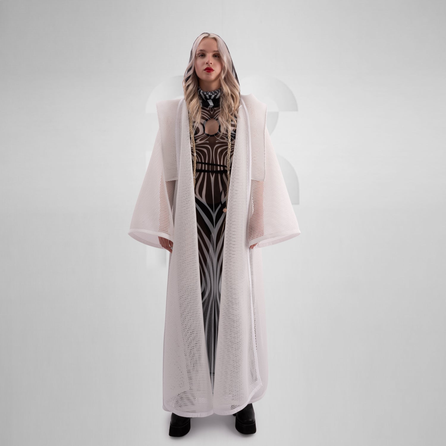 A model wearing a sheer black top with a dramatic white mesh floor-length robe, accessorized with a metallic collar, epitomizing avant-garde fashion and clubwear style, against a neutral gray background.