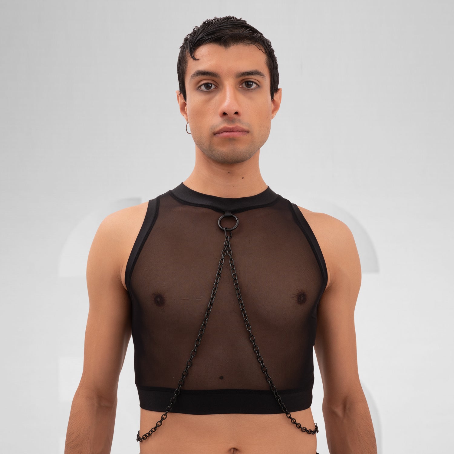 A male model is wearing a black cropped tank top with a sheer mesh fabric covering the torso. The design features a circular metal ring at the center of the chest, with thin black chains extending from the ring to the shoulders and waistband, adding an edgy, modern touch. The background is plain and minimal, emphasizing the bold details of the garment.
