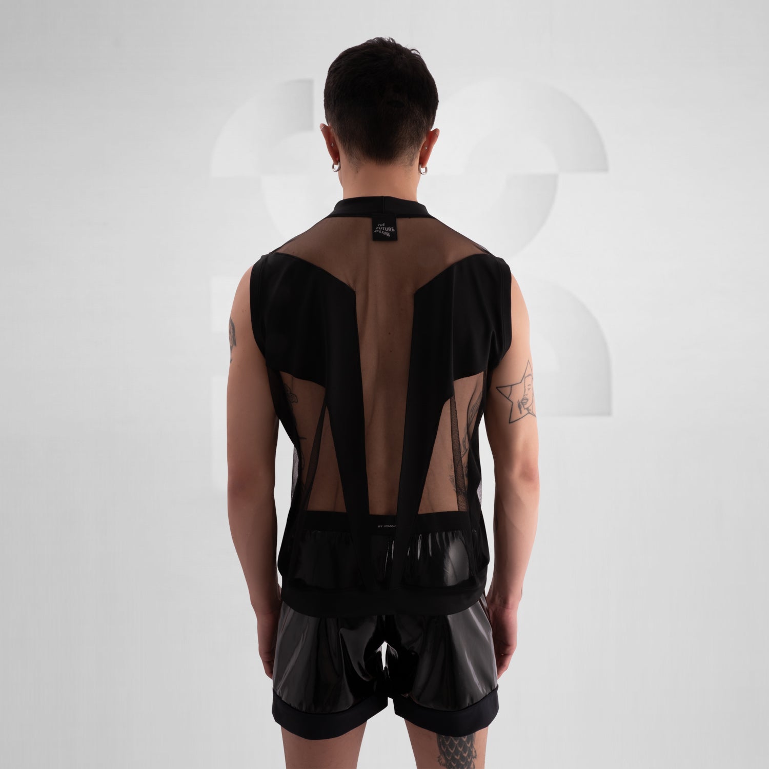  Person wearing a black sleeveless tank top with bold mesh cut-out panels revealing the chest, paired with shiny black vinyl shorts, standing against a neutral background.