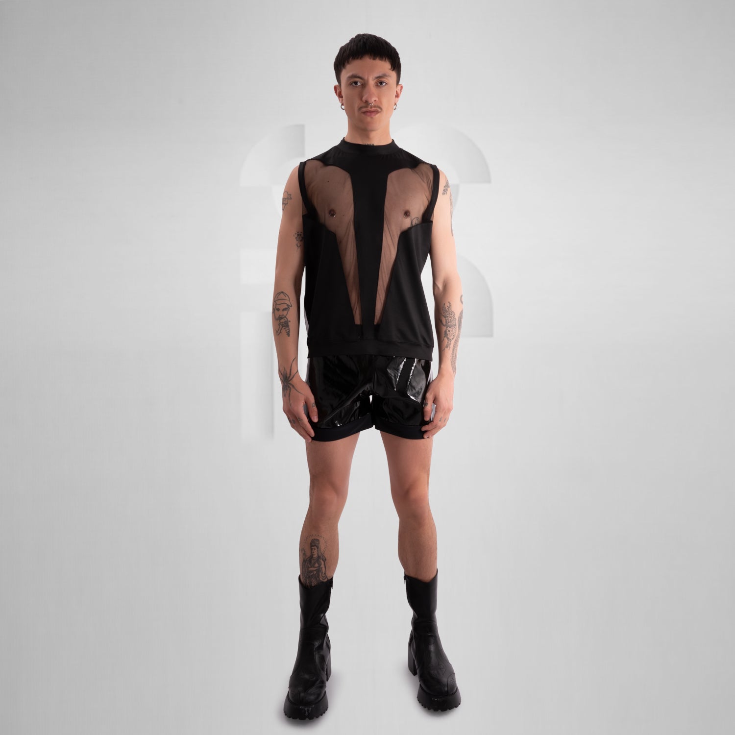 Person wearing a black sleeveless tank top with bold mesh cut-out panels revealing the chest, paired with shiny black vinyl shorts, standing against a neutral background.