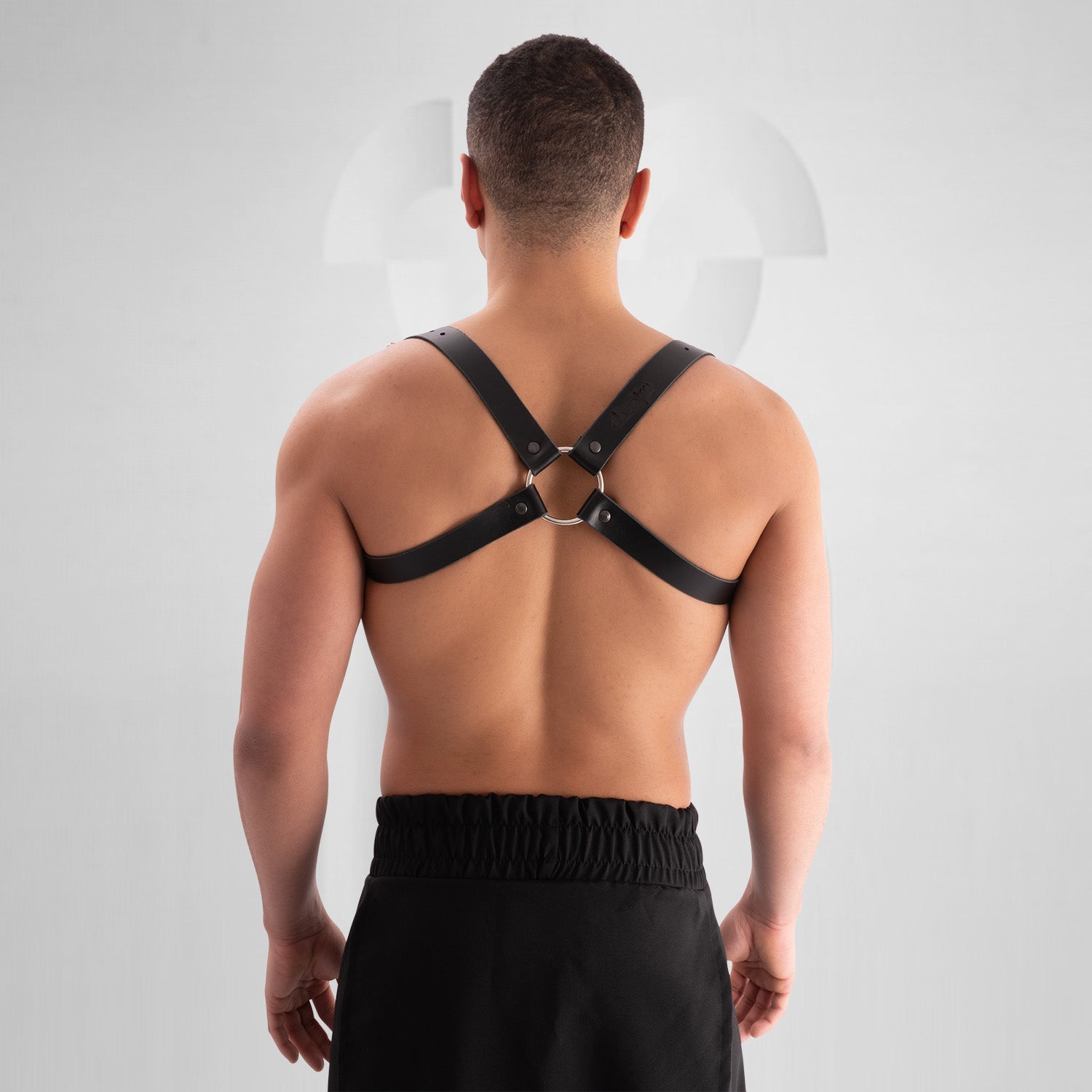 Male model wearing a black leather harness with adjustable buckle straps, featuring a bold crisscross design across the chest. Styled with a black skirt, this avant-garde look is perfect for festival, fetish, and clubwear fashion.