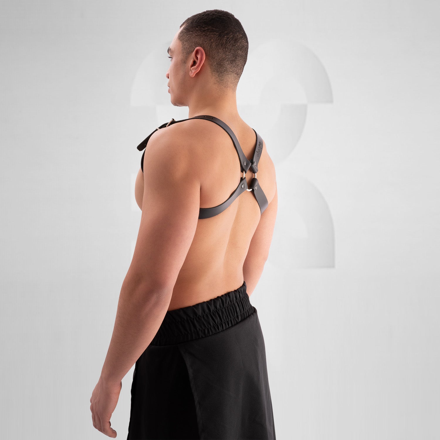 Male model wearing a black leather harness with adjustable buckle straps, featuring a bold crisscross design across the chest. Styled with a black skirt, this avant-garde look is perfect for festival, fetish, and clubwear fashion.