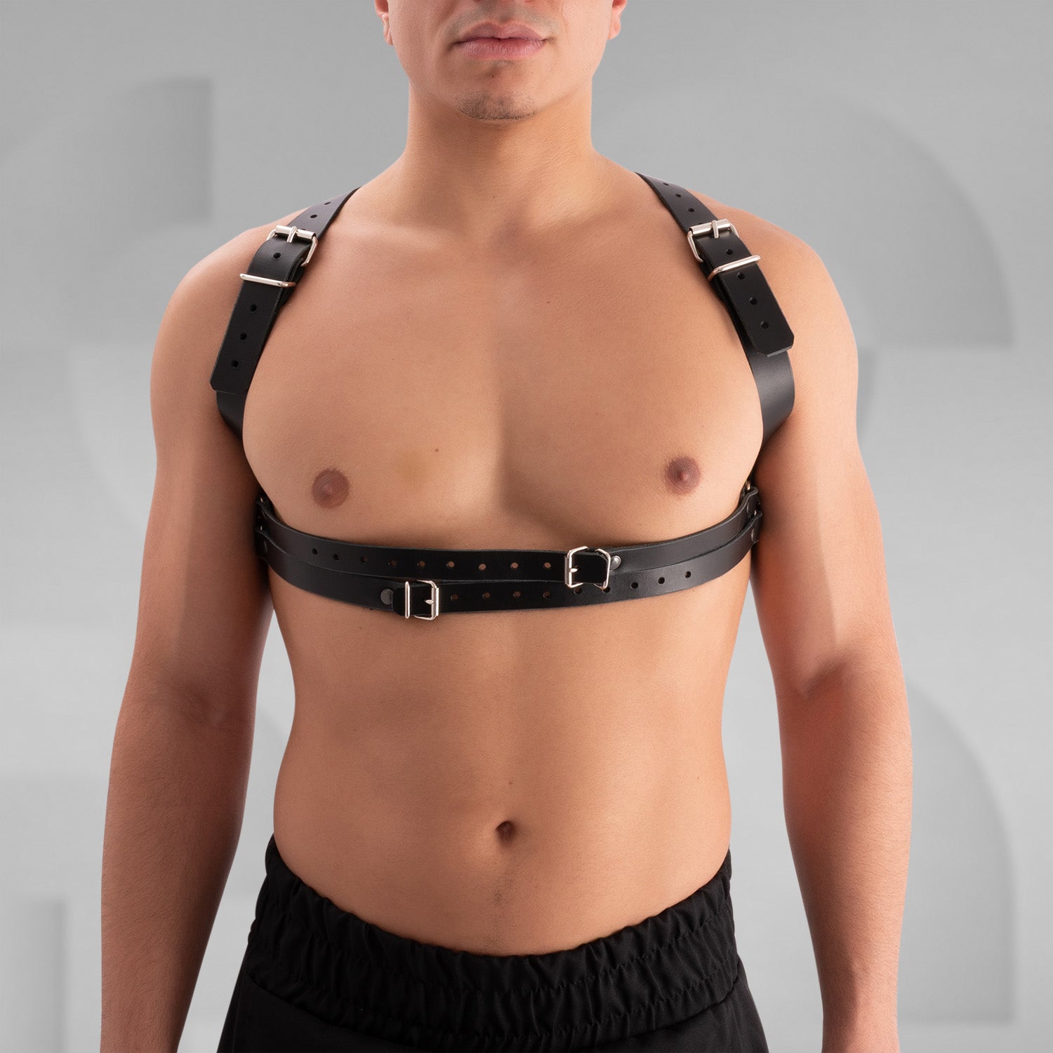 Male model wearing a black leather harness with adjustable buckle straps, featuring a bold crisscross design across the chest. Styled with a black skirt, this avant-garde look is perfect for festival, fetish, and clubwear fashion.