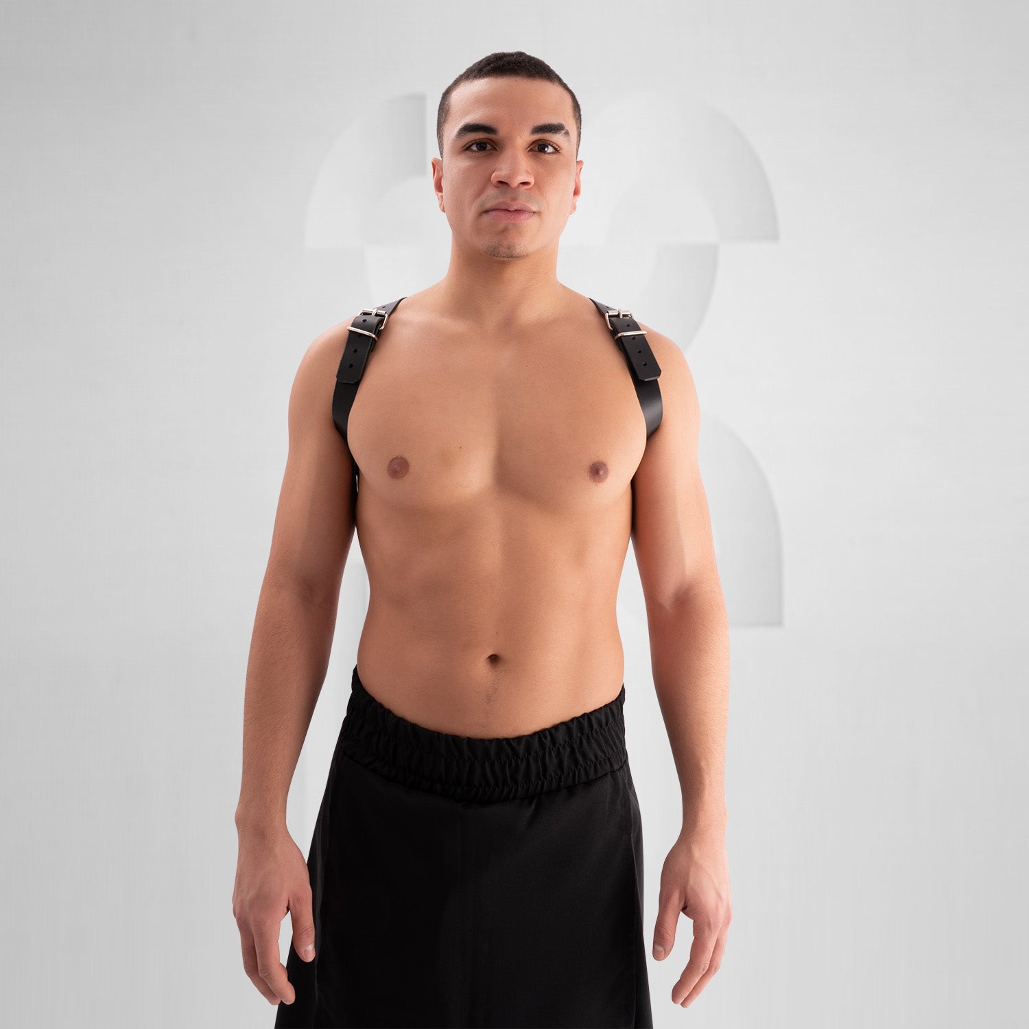 Male model wearing a black leather harness with adjustable buckle straps, featuring a bold crisscross design across the chest. Styled with a black skirt, this avant-garde look is perfect for festival, fetish, and clubwear fashion.
