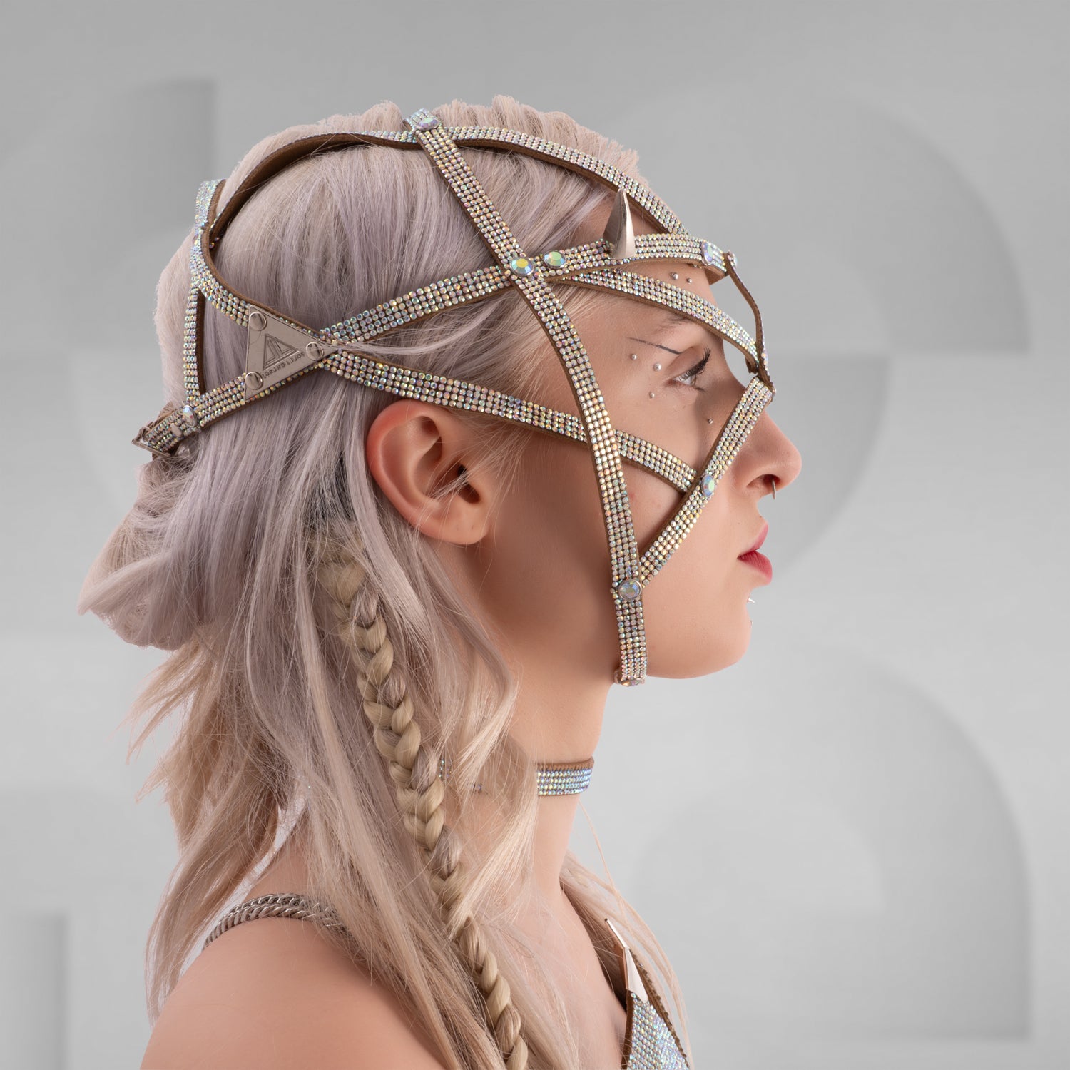 Model wearing a futuristic festival outfit featuring a shimmering silver head harness with angular straps and matching choker, paired with a sculptural, gem-studded metallic bra top that covers the chest in abstract panels. Long platinum-blonde hair is styled in loose waves and braids. Set against a minimal grey background