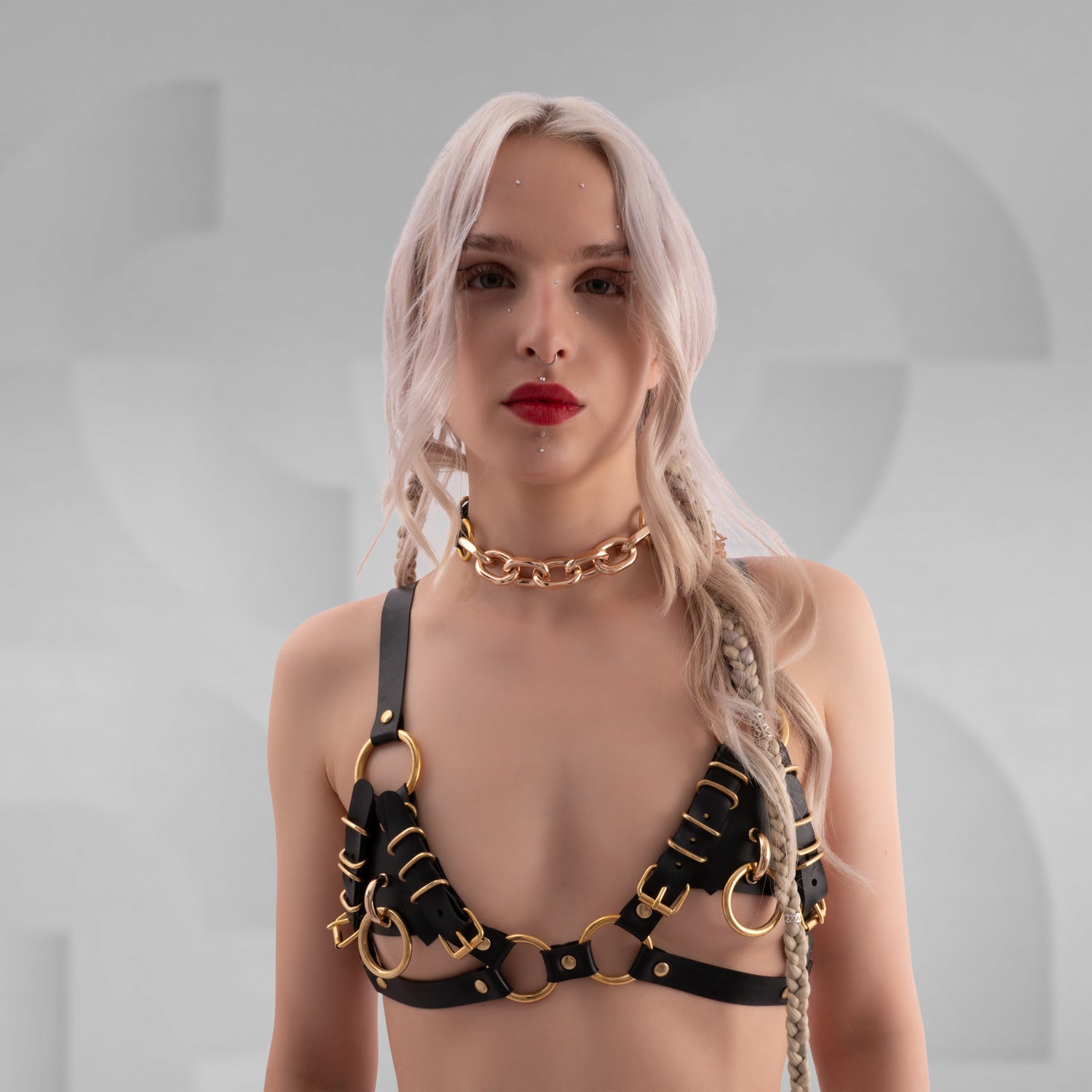 Woman wearing a black leather harness bra with gold metal rings and buckles, paired with a chunky gold chain choker. The outfit features a bondage-inspired aesthetic with intricate strap details and side braids in platinum blonde hair.
