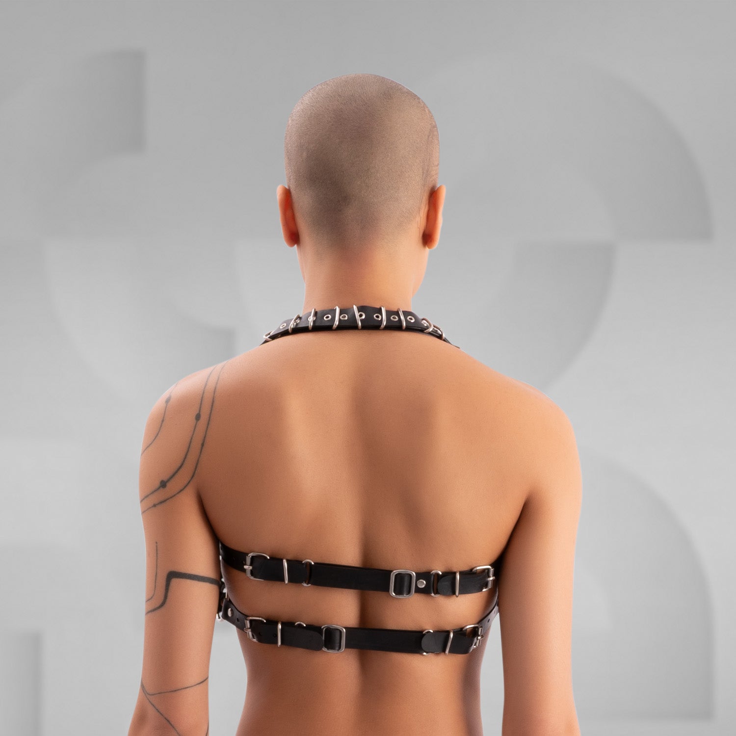 Striking avant-garde rubber bra composed of multiple black straps with silver metal buckles, connected by a large central O-ring. The harness-style design creates a bold, industrial look, worn by a confident model with a shaved head and visible tattoos, standing against a minimalist grey background.