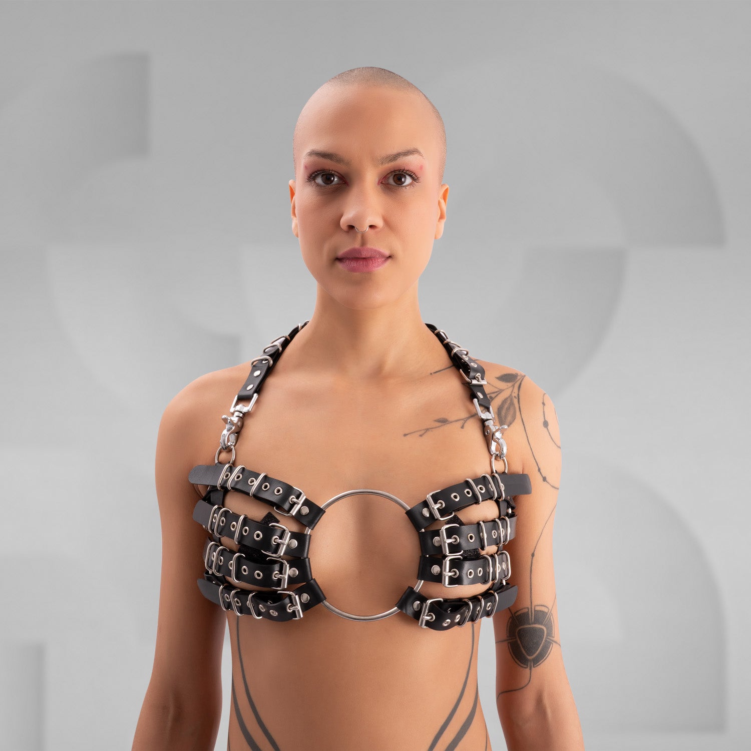 Striking avant-garde rubber bra composed of multiple black straps with silver metal buckles, connected by a large central O-ring. The harness-style design creates a bold, industrial look, worn by a confident model with a shaved head and visible tattoos, standing against a minimalist grey background.