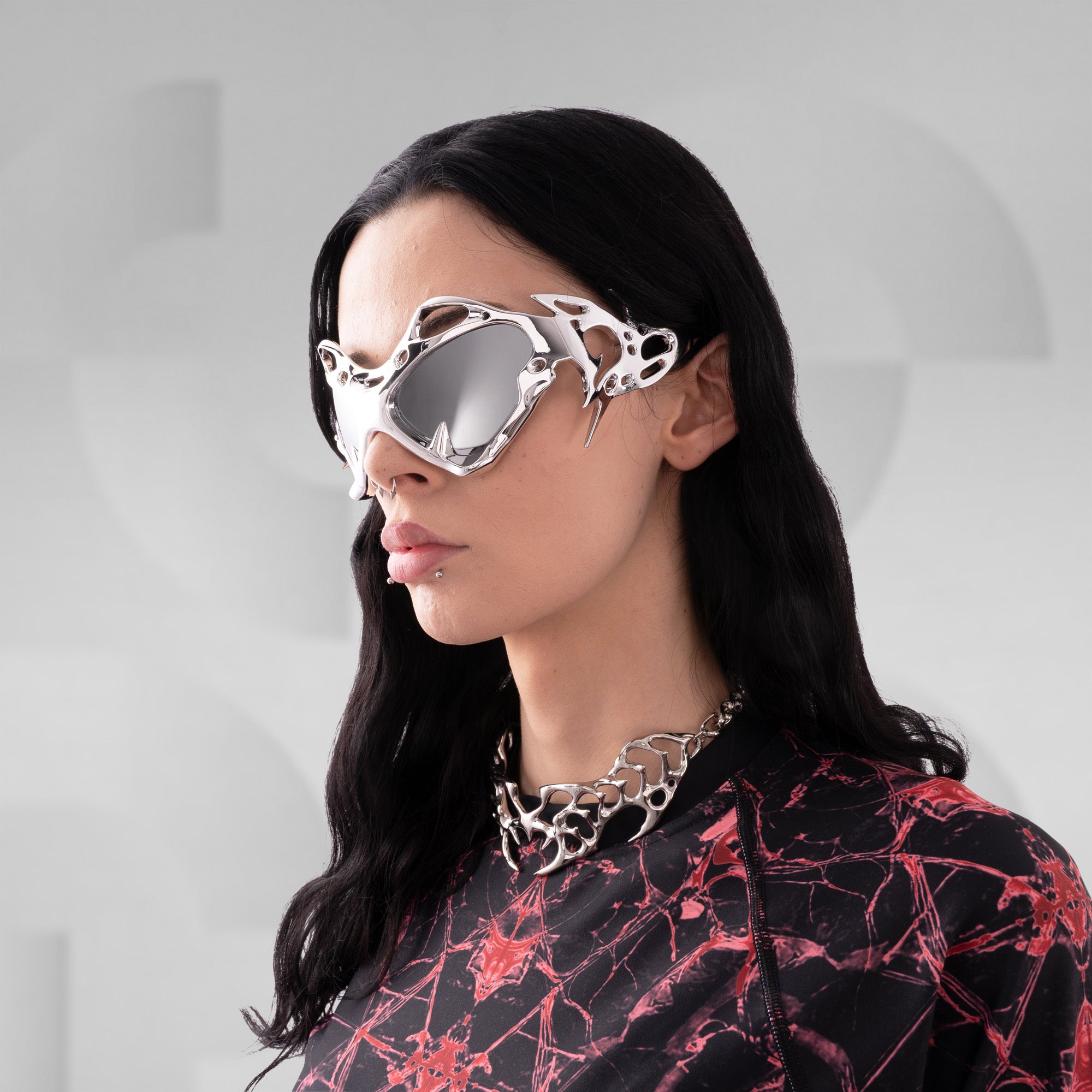 Person wearing futuristic sunglasses and a black and red patterned outfit against a gray background