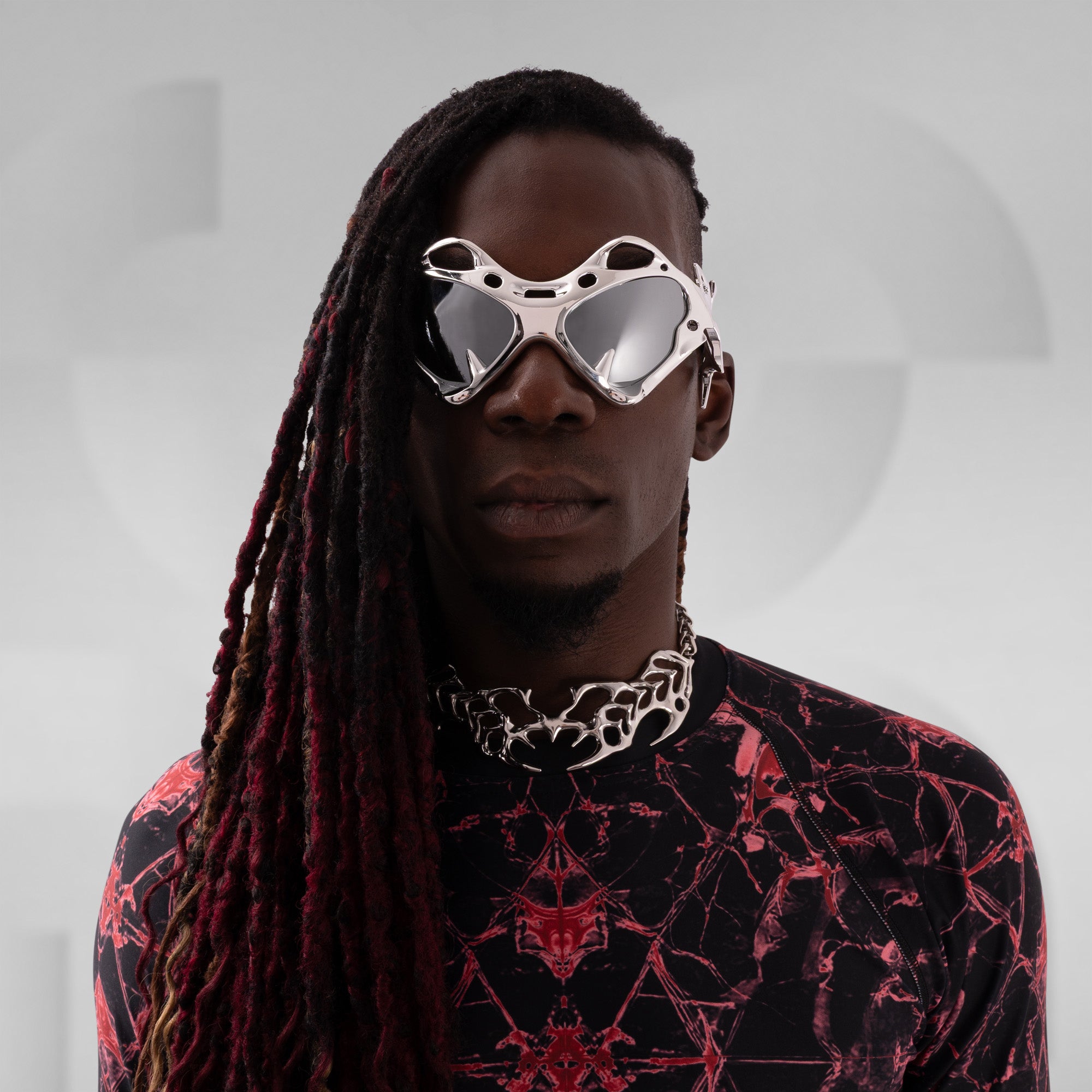 Person wearing futuristic sunglasses and a black and red patterned outfit on a gray background