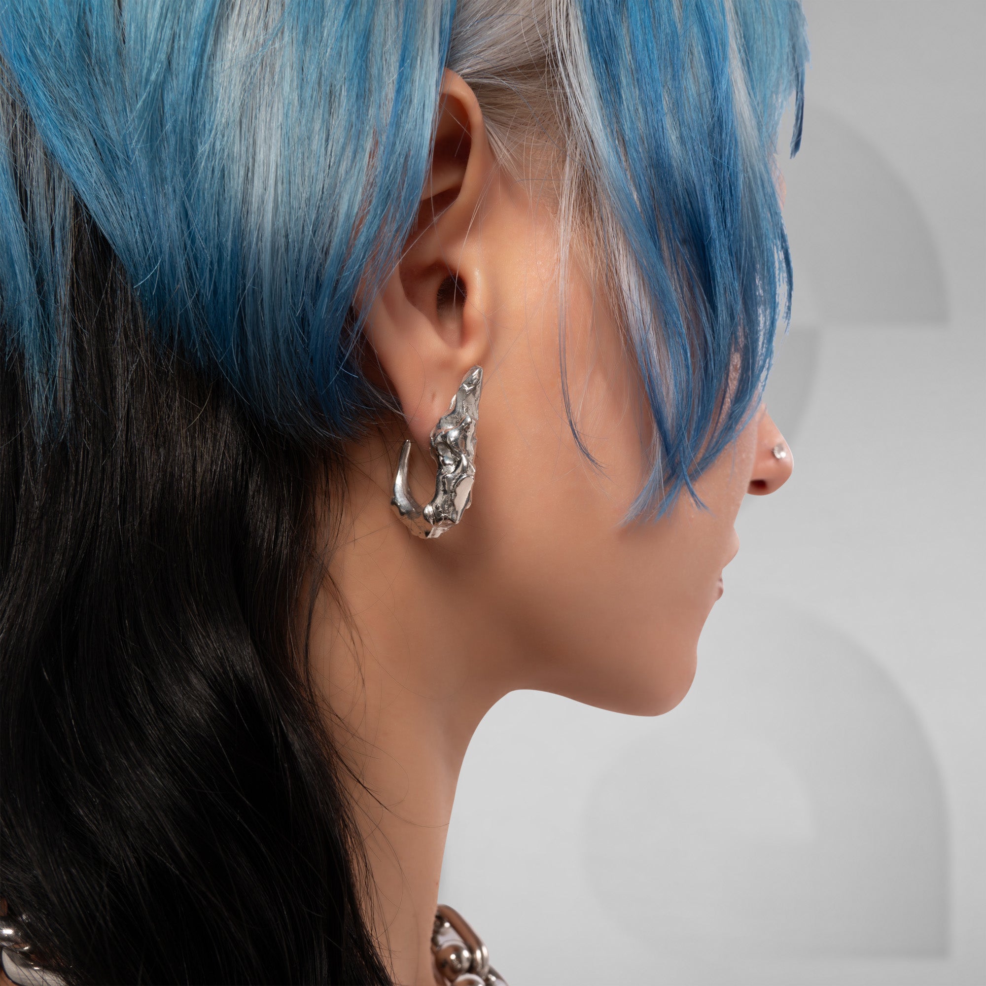 A close-up side profile of a person with blue and black hair wearing a textured silver huggie hoop earring.