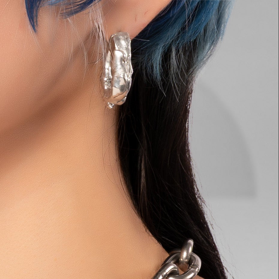 A close-up of a model wearing a chunky silver chain choker necklace, with black hair, blue highlights, and visible chest tattoos against a soft gray background.