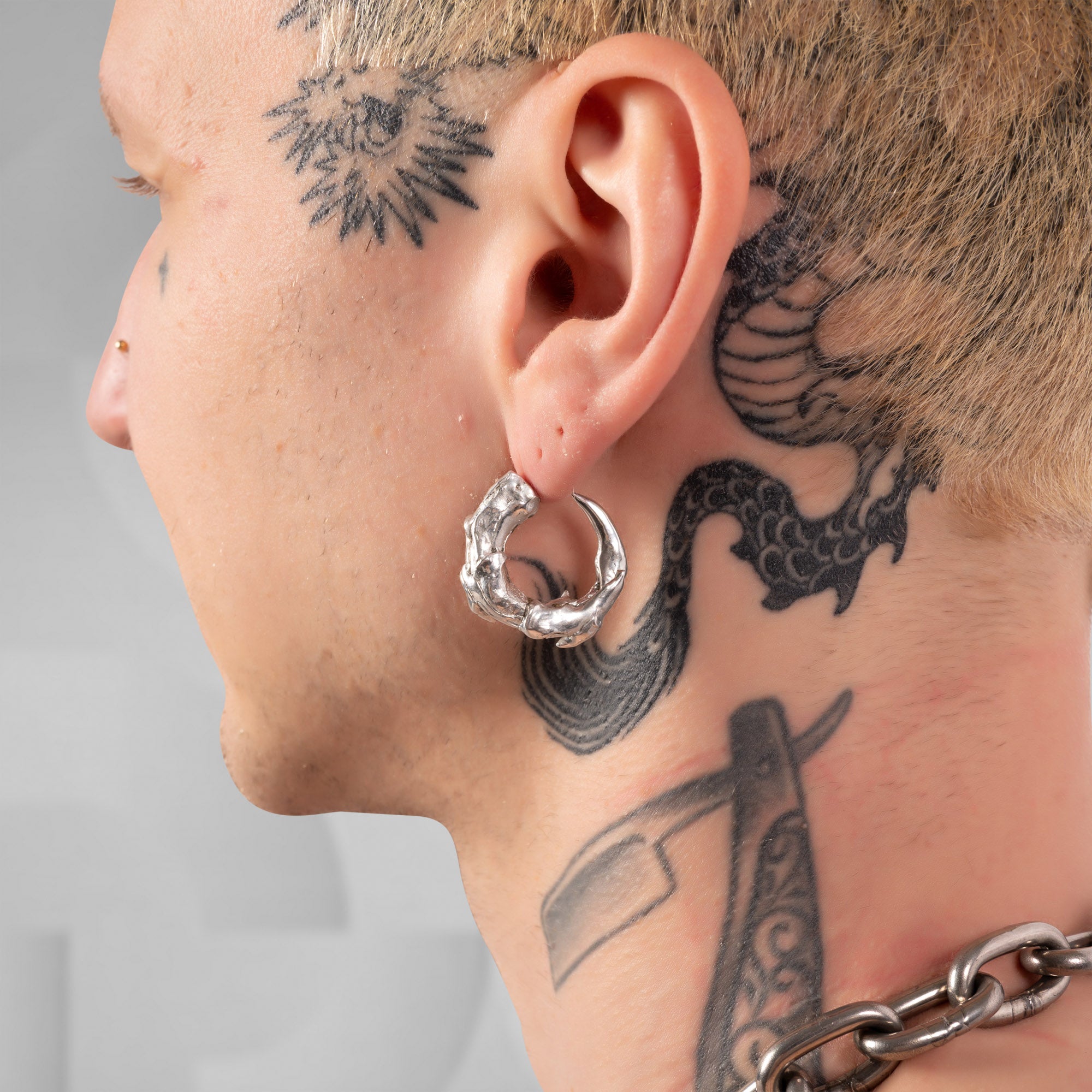 A close-up side profile of a person's head and neck, showing a textured silver hoop earring worn in the ear. The person has visible tattoos on the head, neck, and side of the face, and a silver chain is visible at the bottom right.