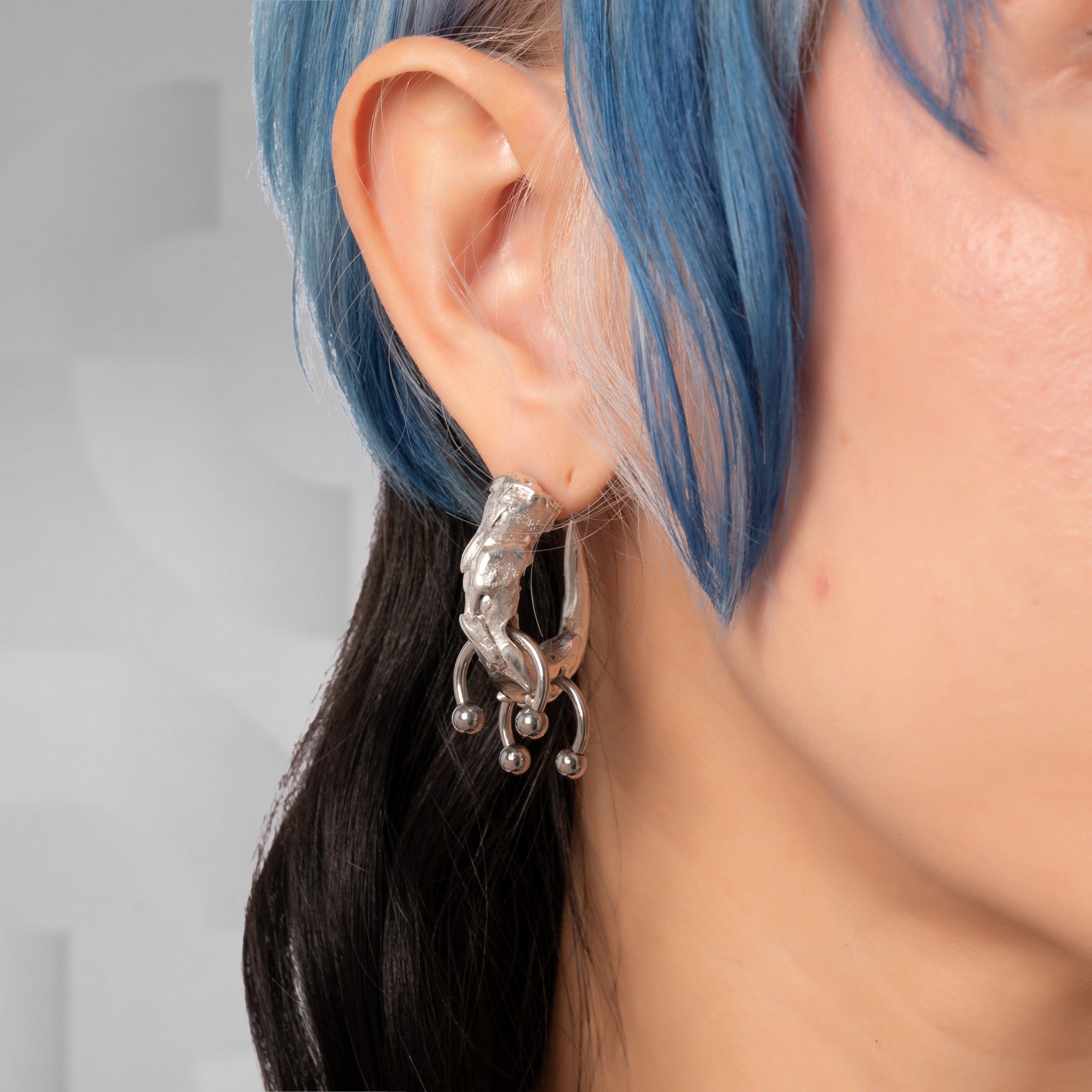 A close-up of a person's ear wearing a chunky, textured silver huggie hoop earring with dangling beaded loops, set against a soft gray background.
