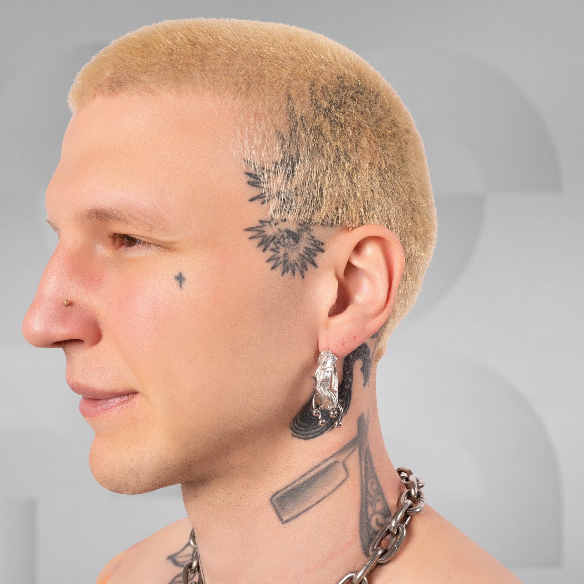 Close-up side profile of a tattooed man with a short blond haircut, black floral tattoo on the side of his head, a small cross tattoo on his cheek, and a chunky silver hoop earring.