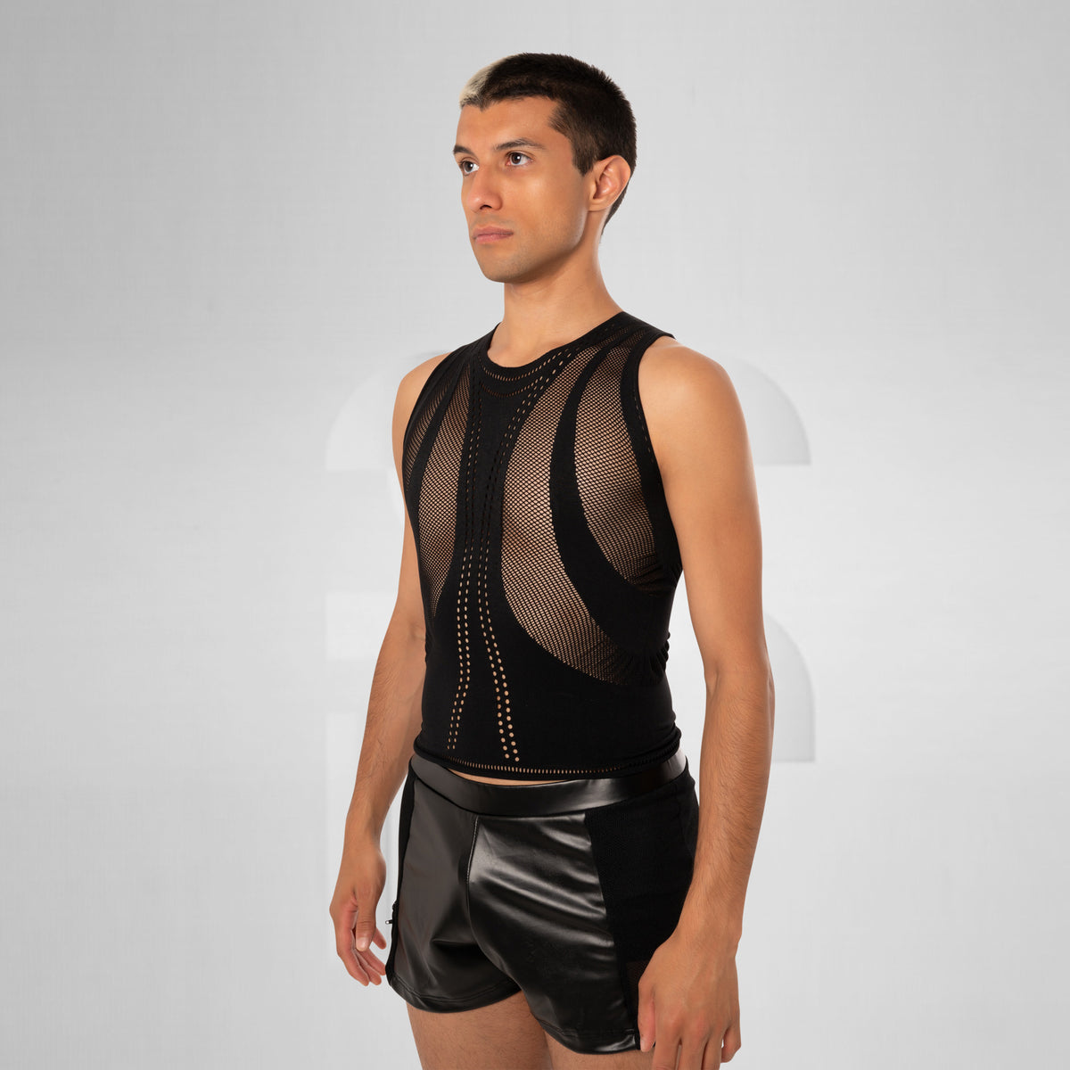 A man modeling a black sleeveless mesh bodysuit with structured panels and black faux-leather shorts against a light gray studio background.