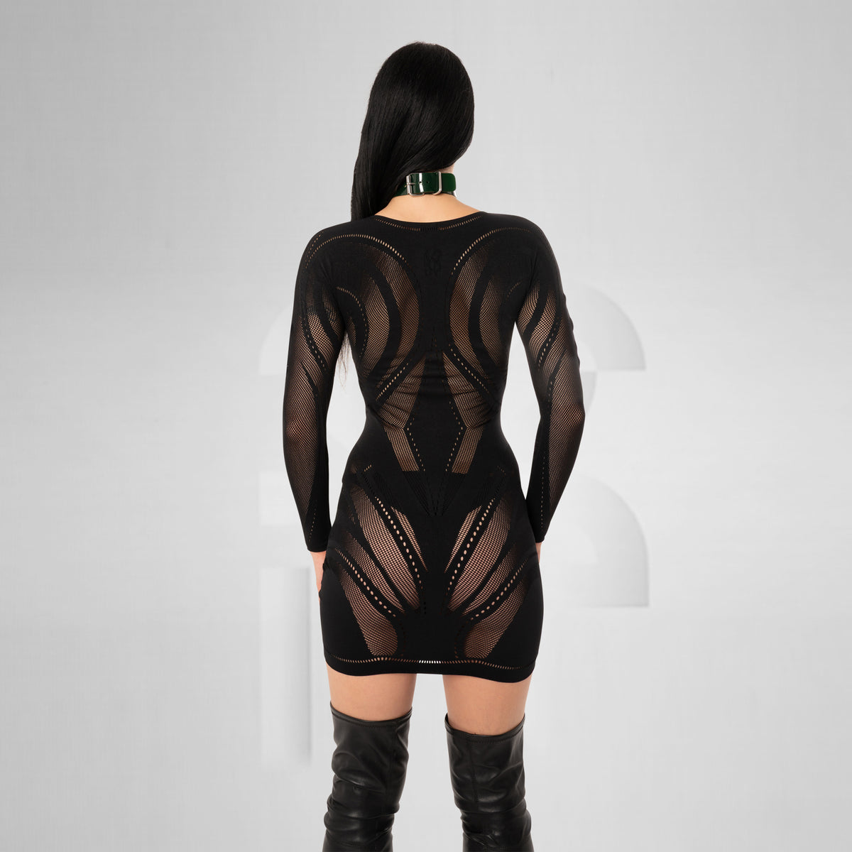 A woman viewed from the back wearing a black sheer long-sleeve mini dress with geometric cutout patterns and black thigh-high boots against a light gray studio background.