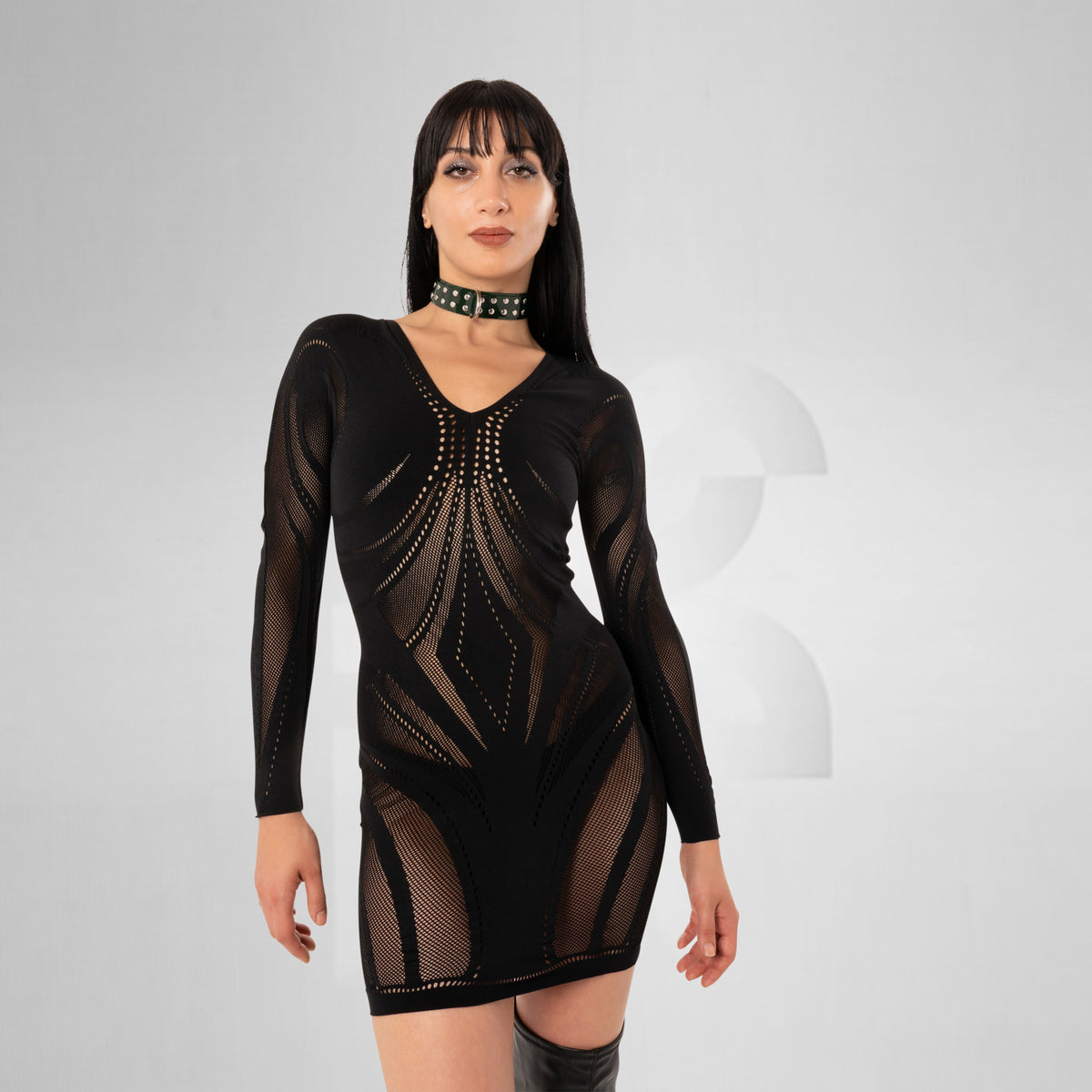 A woman modeling a black long-sleeve mini dress with sheer mesh panels and geometric cutout details, paired with a black studded choker, against a light gray studio background.