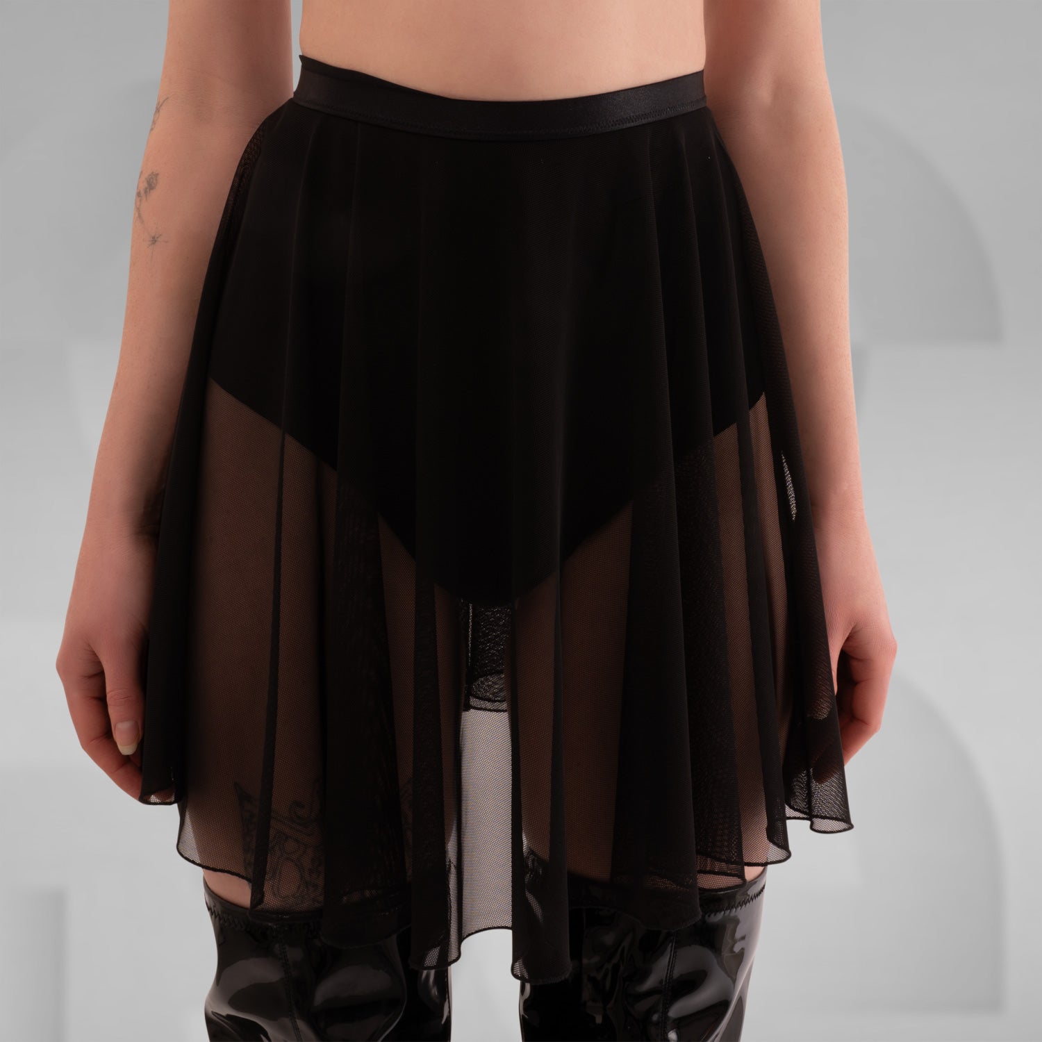 The a close-up of a sheer black skirt with a flowing, layered design. It is paired with a darker underskirt, highlighting the contrast between the two fabrics. The model's arms, adorned with tattoos, are visible in the frame, adding an artistic element to the image. The neutral background emphasizes the textures and details of the skirt and the overall look.