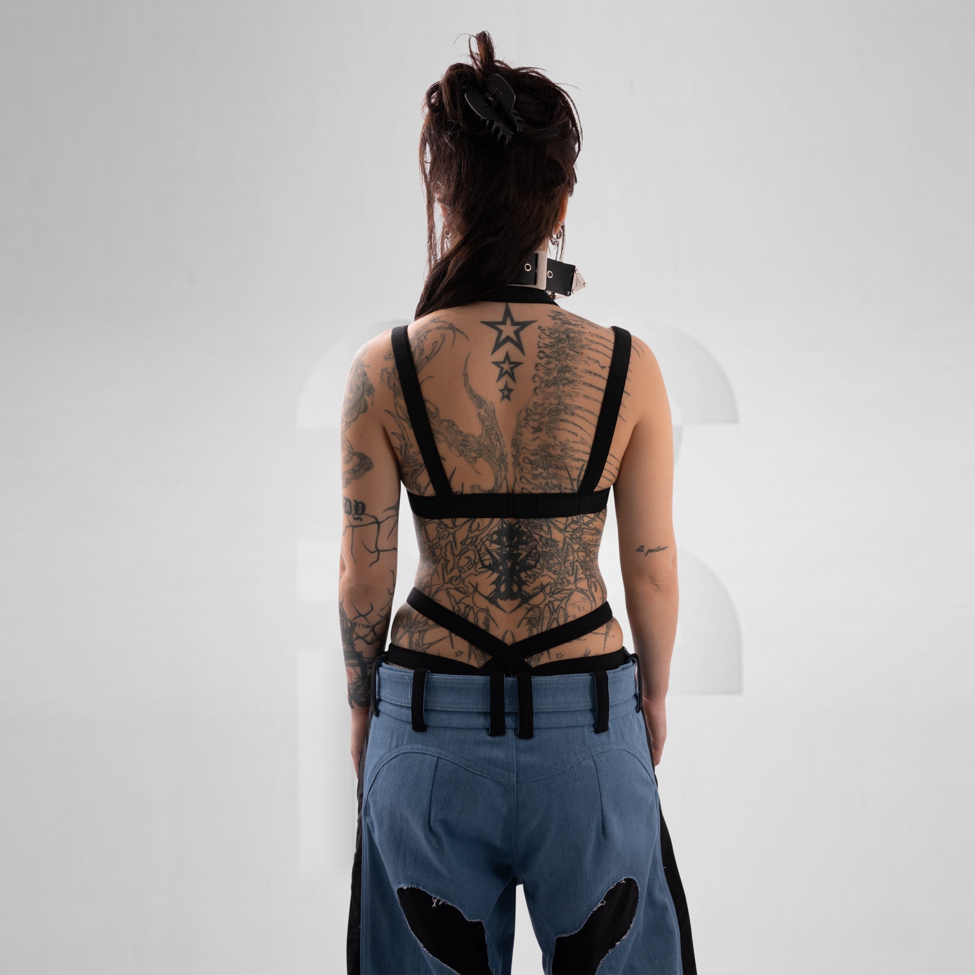 Person wearing a black harness with tattoos on a plain background