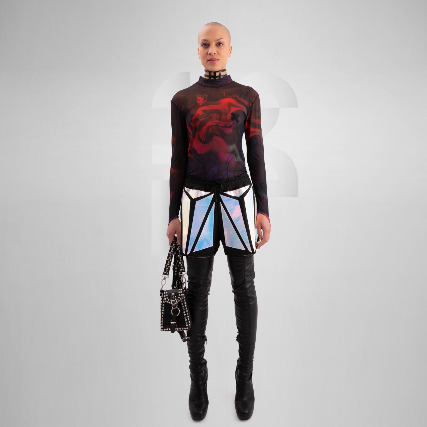Person wearing a black studded leather harness bag with silver hardware and rings, styled over a sheer mesh top featuring vibrant red and purple abstract artwork. The edgy, fashion-forward look is completed with holographic geometric-patterned pants, captured from behind against a minimalist grey background.