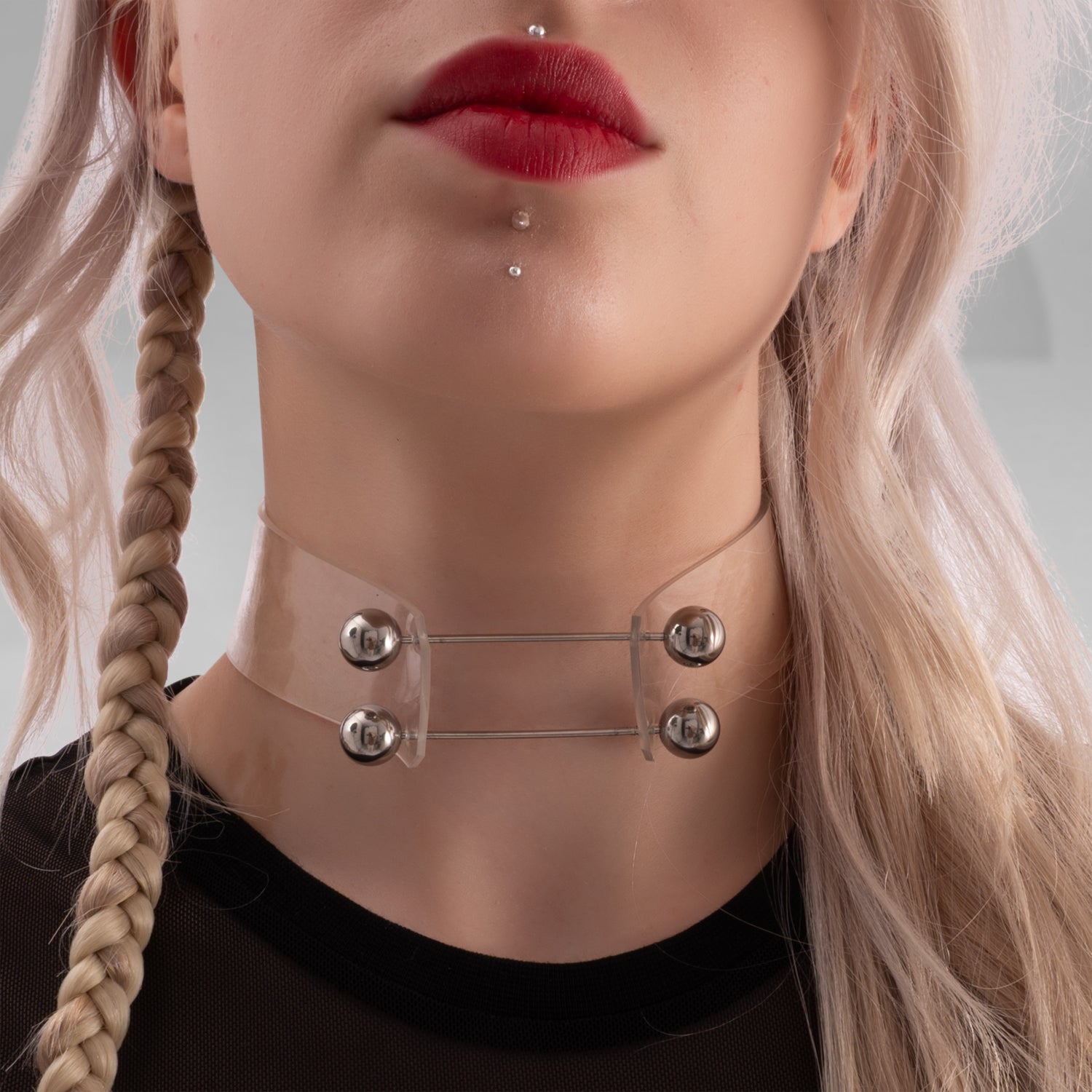 The image showcases a woman with platinum blonde hair styled in loose waves and a unique collection of jewelry. She wears a clear, futuristic choker with metal spheres, a septum piercing, and a labret stud. Her makeup includes bold red lipstick and artistic eye makeup with fine black lines and small studs near her eyes. The overall look is avant-garde and edgy, perfect for making a statement.