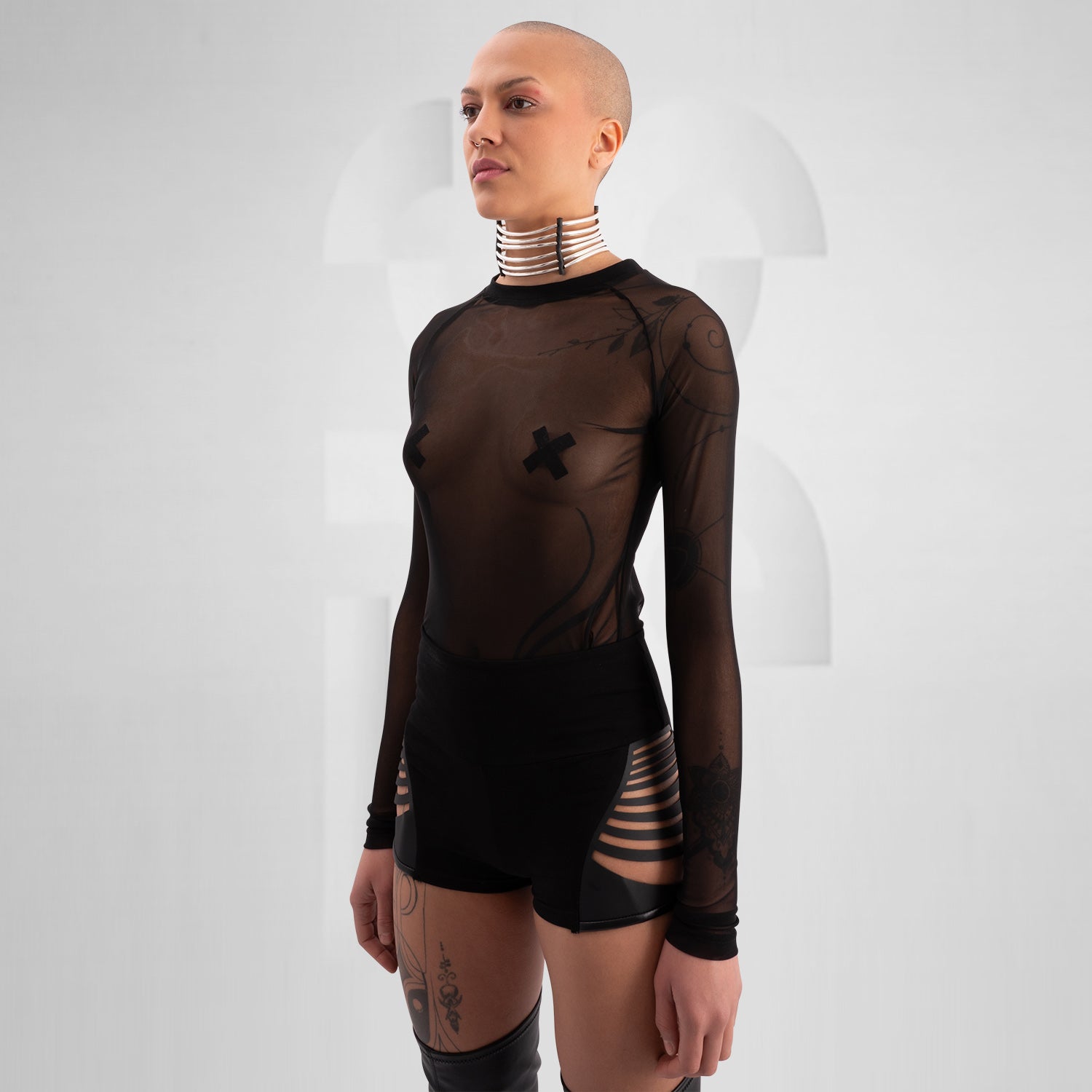 Model wearing a bold, futuristic black sheer long-sleeve top with a high neckline, paired with black cut-out shorts. The look is completed with a statement metallic choker and nipple pasties, creating an edgy, avant-garde aesthetic. Perfect for techno, festival, and alternative fashion