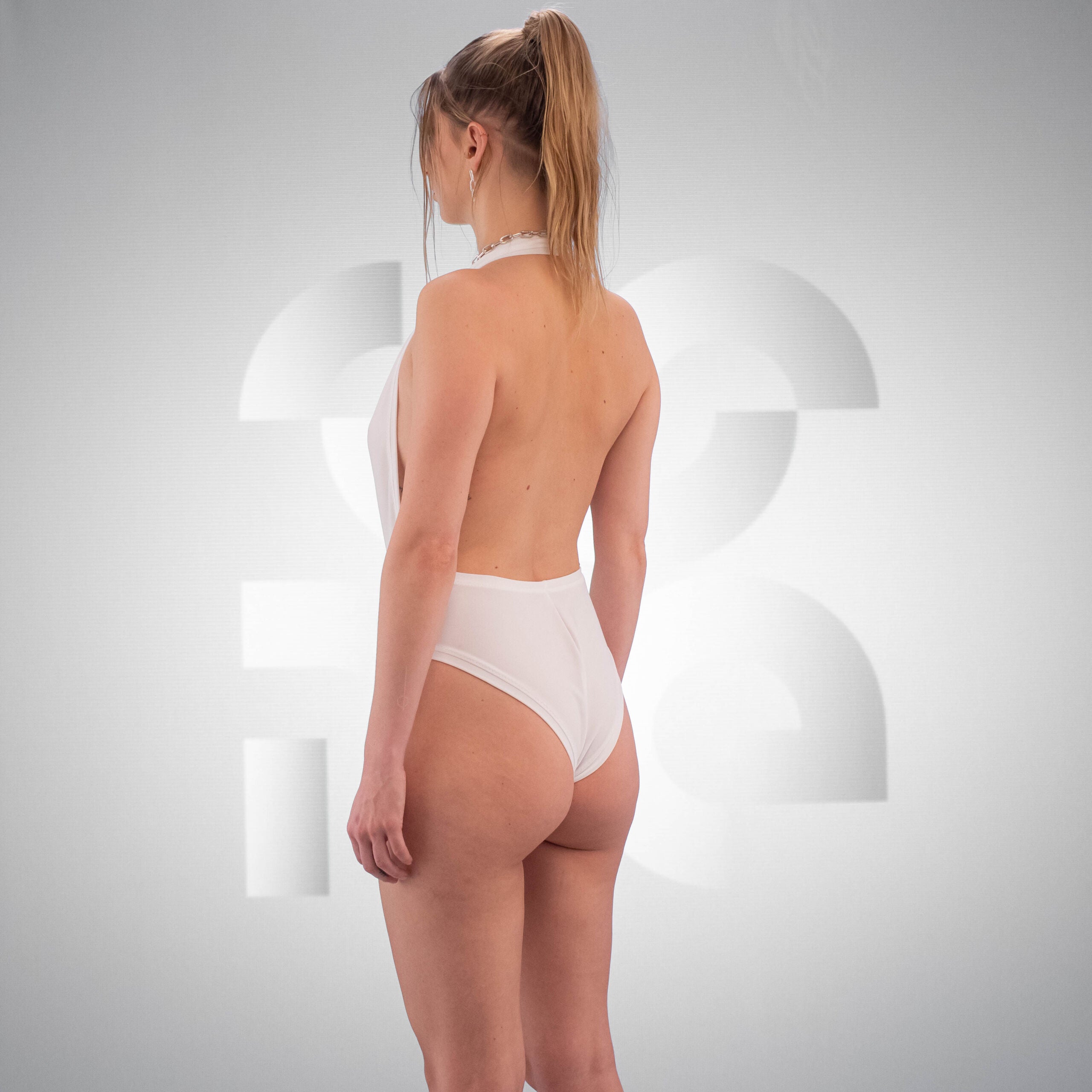 The wearing a sleek, white one-piece swimsuit with a deep V neckline and high cut legs. Their hair is tied back in a high ponytail, and they have a confident expression. Minimal jewelry, including a simple necklace, complements the look.