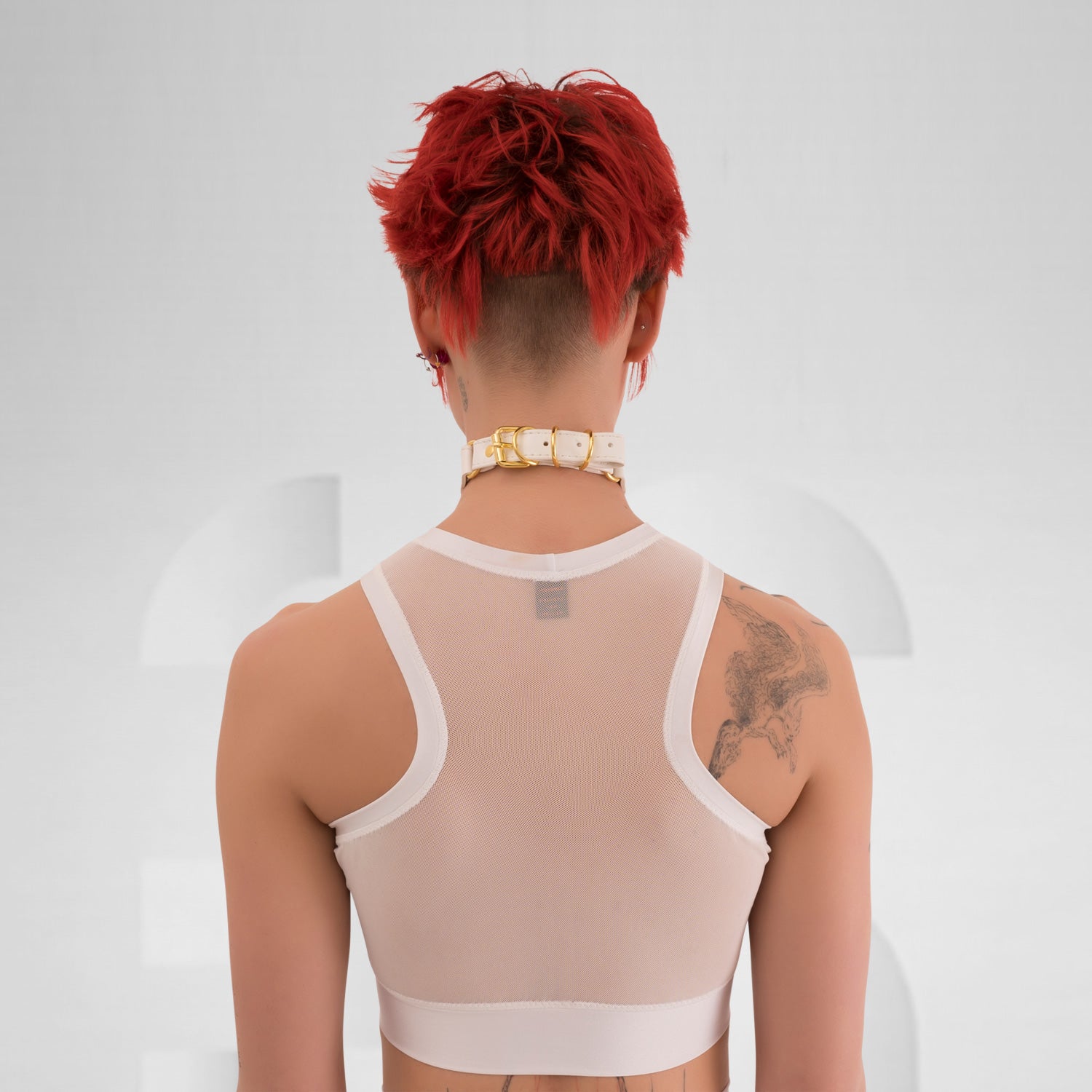 The side profile of a model wearing a translucent white top with a modern, sporty design. The top is complemented by a choker that has a gold detail, adding an edgy touch. The model has short, vibrant red hair and dramatic makeup, enhancing their bold look. The neutral background focuses attention on the outfit and the striking features of the model.