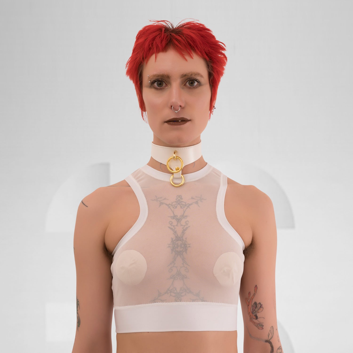 The side profile of a model wearing a translucent white top with a modern, sporty design. The top is complemented by a choker that has a gold detail, adding an edgy touch. The model has short, vibrant red hair and dramatic makeup, enhancing their bold look. The neutral background focuses attention on the outfit and the striking features of the model.