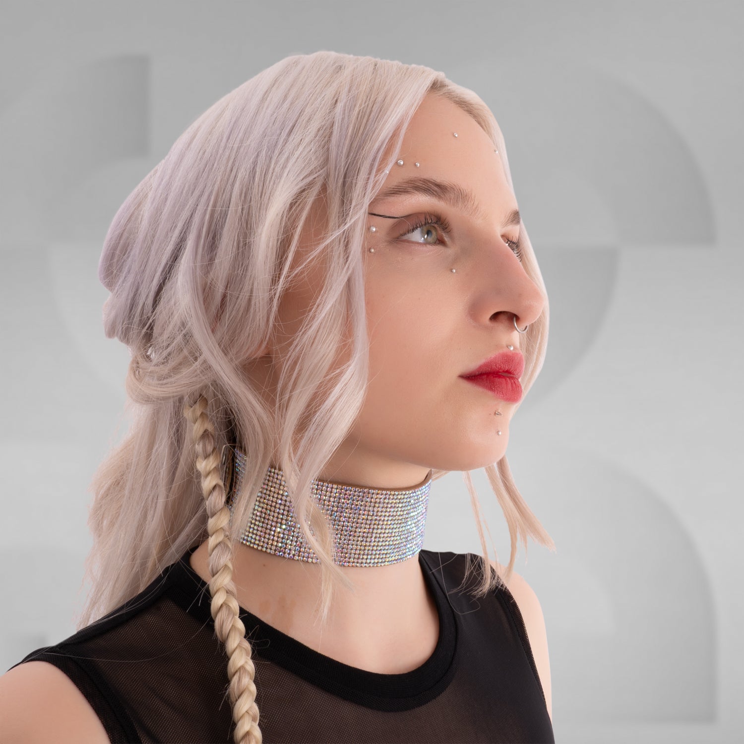 This close-up image highlights a woman with a daring, edgy style. She has platinum blonde hair styled in loose waves with a braid on one side. She's wearing a sparkling, multi-colored rhinestone choker around her neck, which adds a glamorous, bold statement to her look. Her makeup features vibrant red lipstick matching her accessories, and she has multiple facial piercings, including a septum ring and dermal piercings below her lips, emphasizing her alternative, fashion-forward aesthetic.