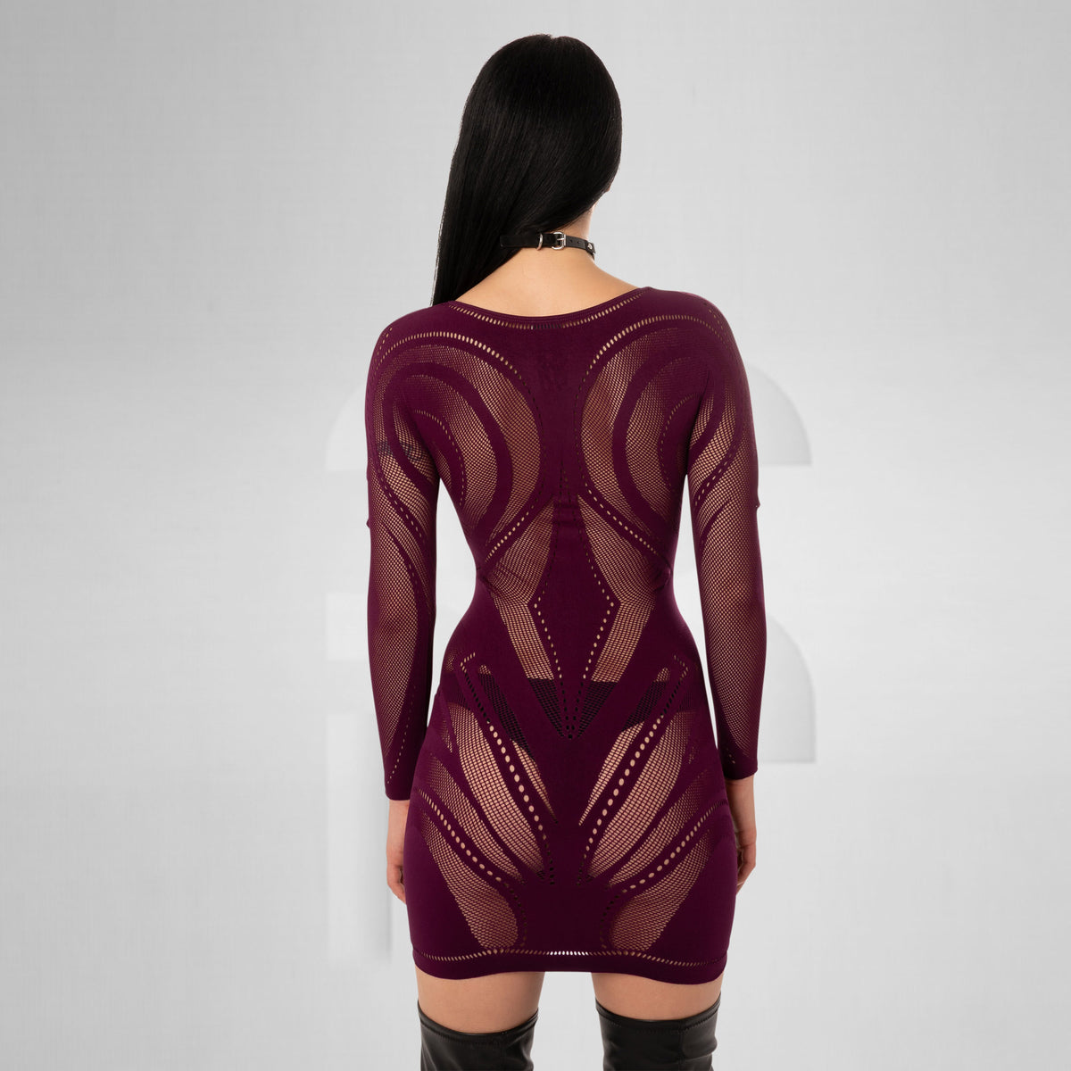 A woman viewed from the back wearing a long-sleeve, bodycon mini dress made of sheer burgundy mesh with geometric cutout patterns. She is standing against a light gray studio background.